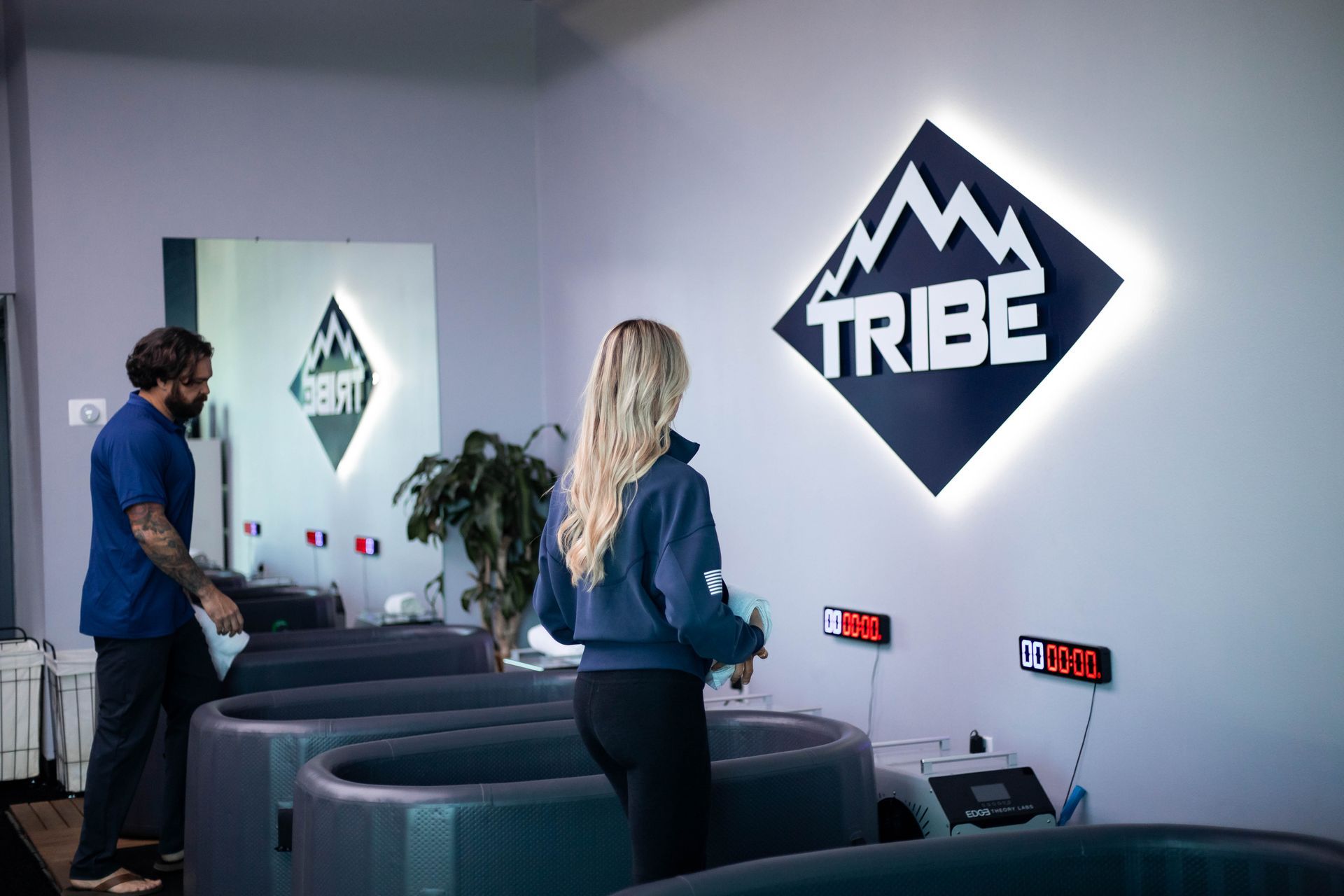 Two people at a "Tribe" health center with ice baths. A logo is on the wall.