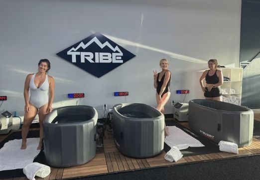 Three women in swimsuits pose near cold plunge tubs, under a "TRIBE" sign.