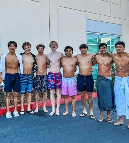 Group of eight young men in swimwear, smiling, standing together outdoors near a building.