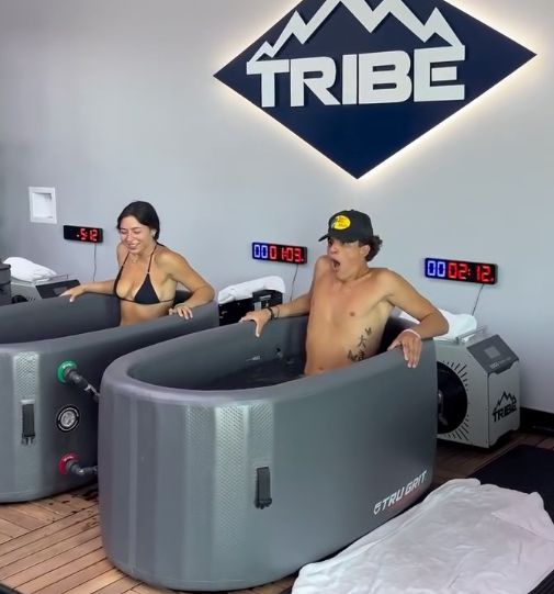 Two people in cold plunge tubs at Tribe. Woman grimaces, man looks shocked. White room, blue logo.