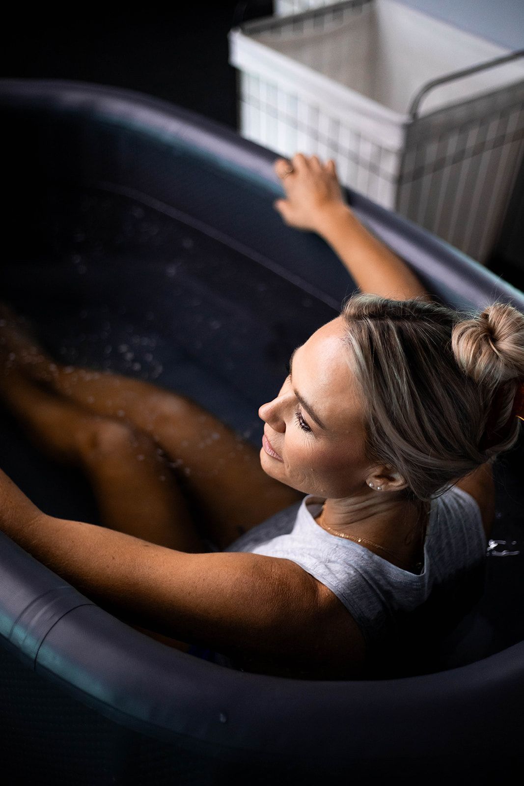 Woman in a dark tub, head back, eyes closed, blonde hair up.
