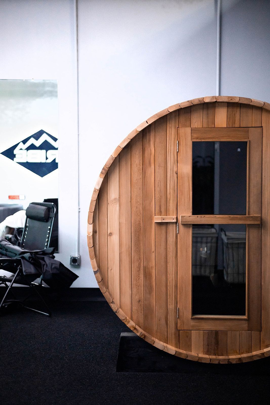 Wooden barrel sauna with a glass window, next to an office wall with logo and chair.