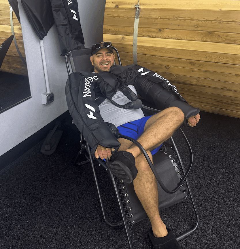 Man in compression sleeves smiling while reclined in a chair. Gym setting.