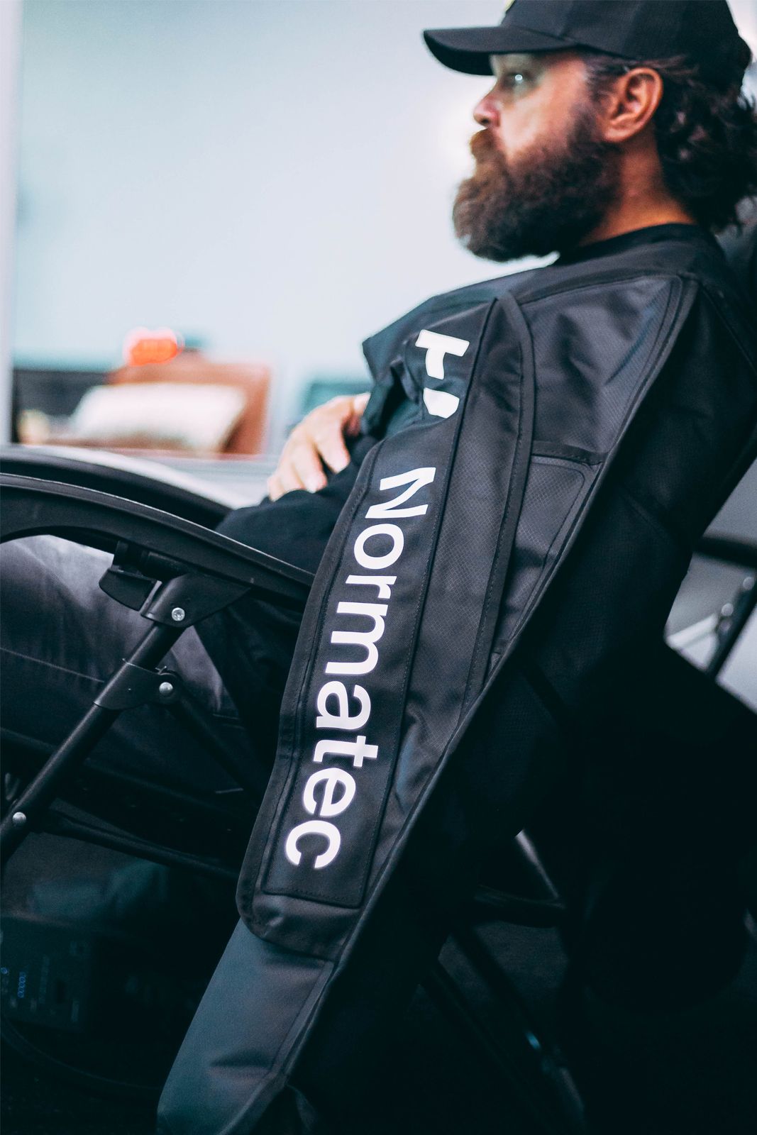 Man in chair with a Normatec sleeve on his arm. Wearing a black hat and shirt, with a beard.