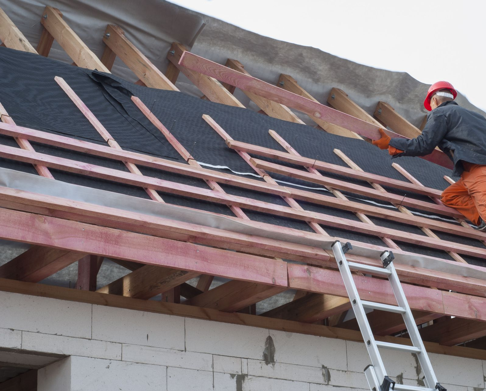 Roofer on ladder installing black underlayment on wooden roof frame.