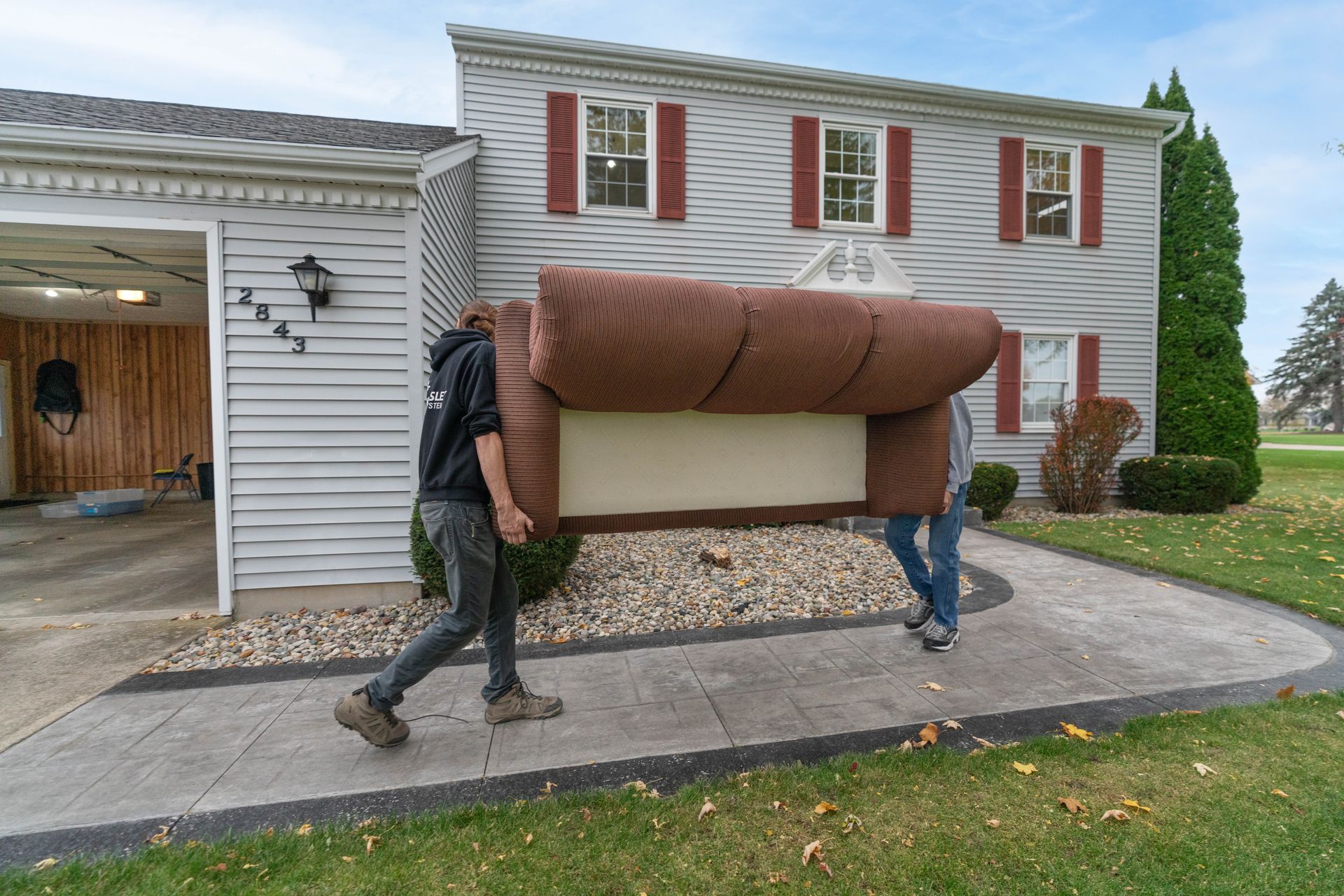 Two people carrying a brown sofa up a driveway beside a white house and garage.