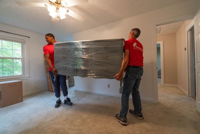 Two movers carrying a wrapped couch through a carpeted room in a house.