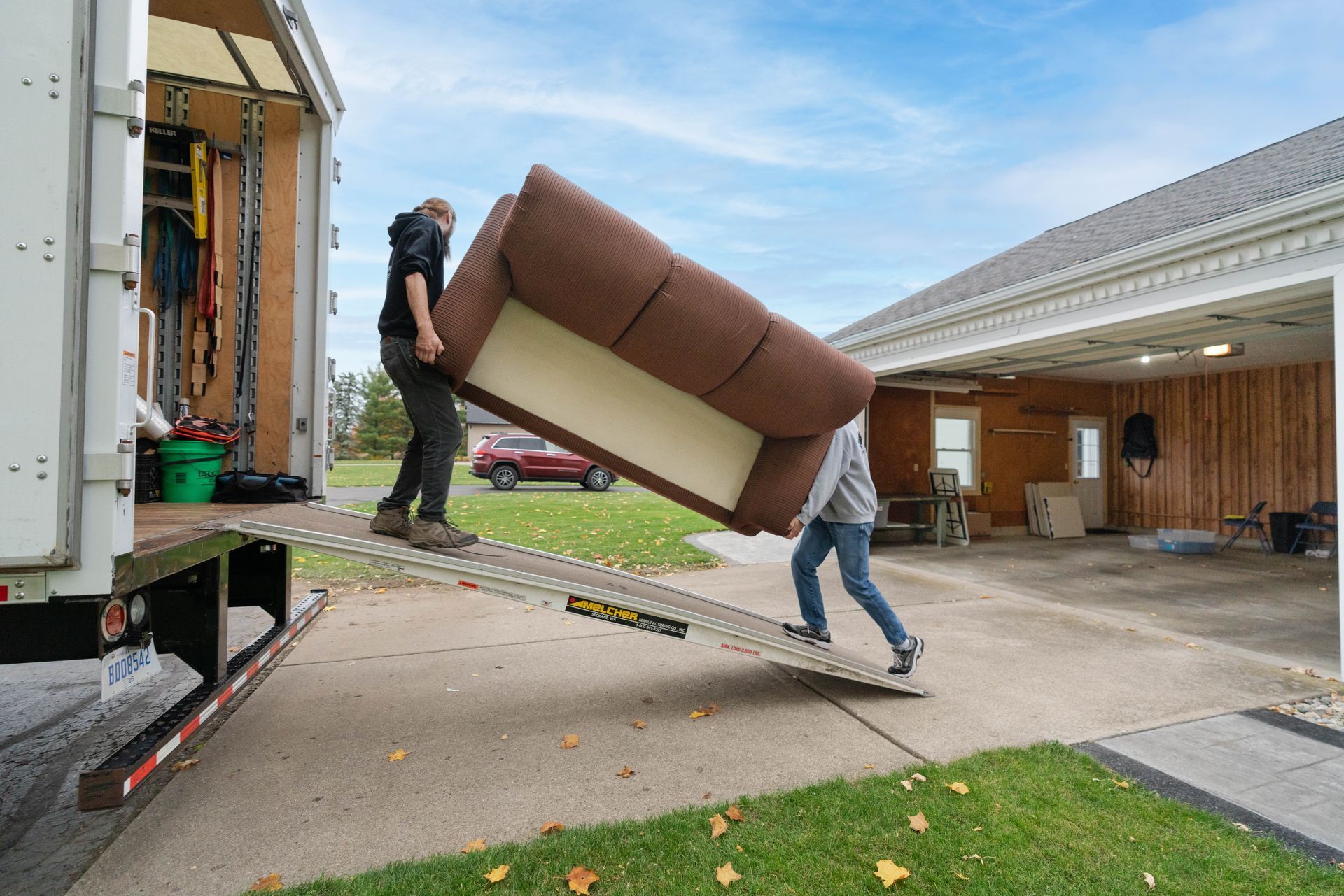 Two movers carrying a brown couch from a truck into a suburban driveway