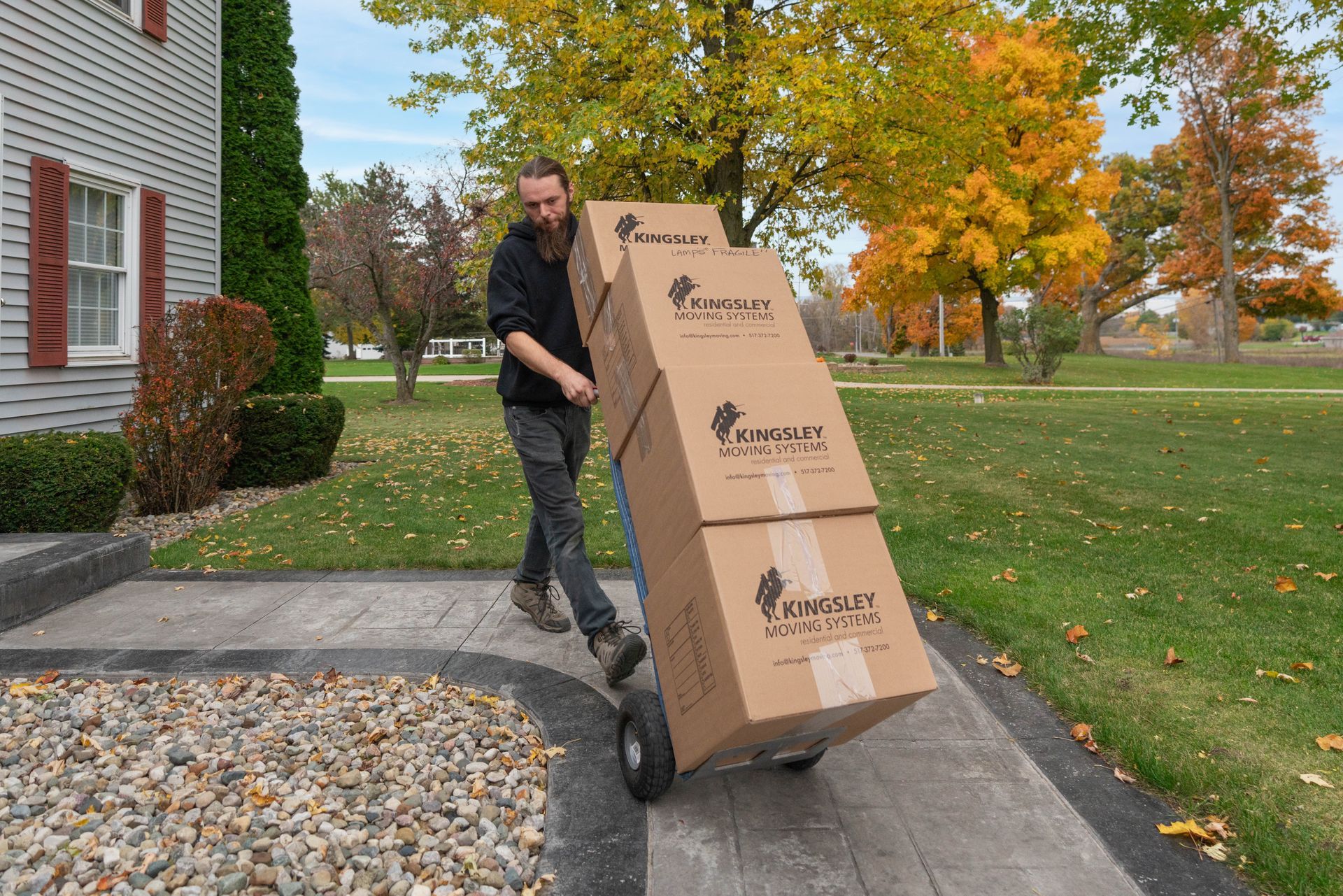 Man pushing a tall cardboard box up a walkway beside a house, with autumn trees in the background