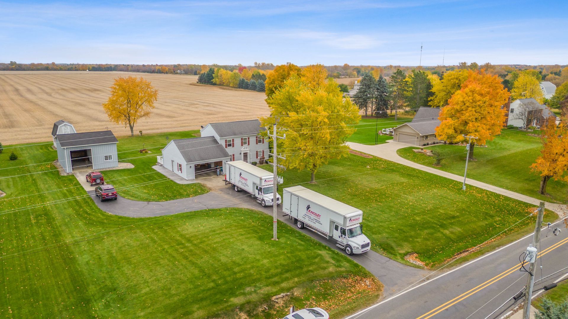 Rural farmhouse and white RV beside a road, surrounded by green fields and autumn trees
