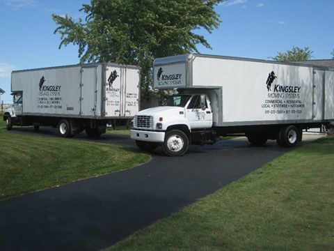 Two white moving trucks parked on a driveway beside a green lawn under a tree.