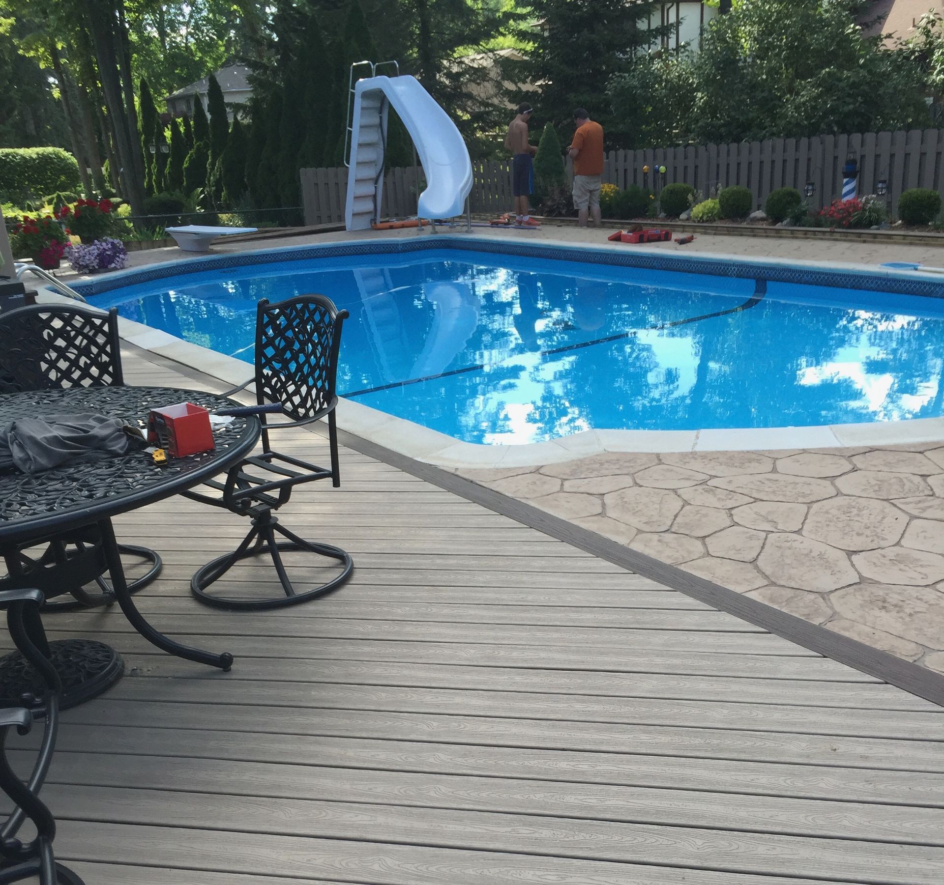 Backyard pool with a slide, surrounded by a deck and stone patio, with people working.