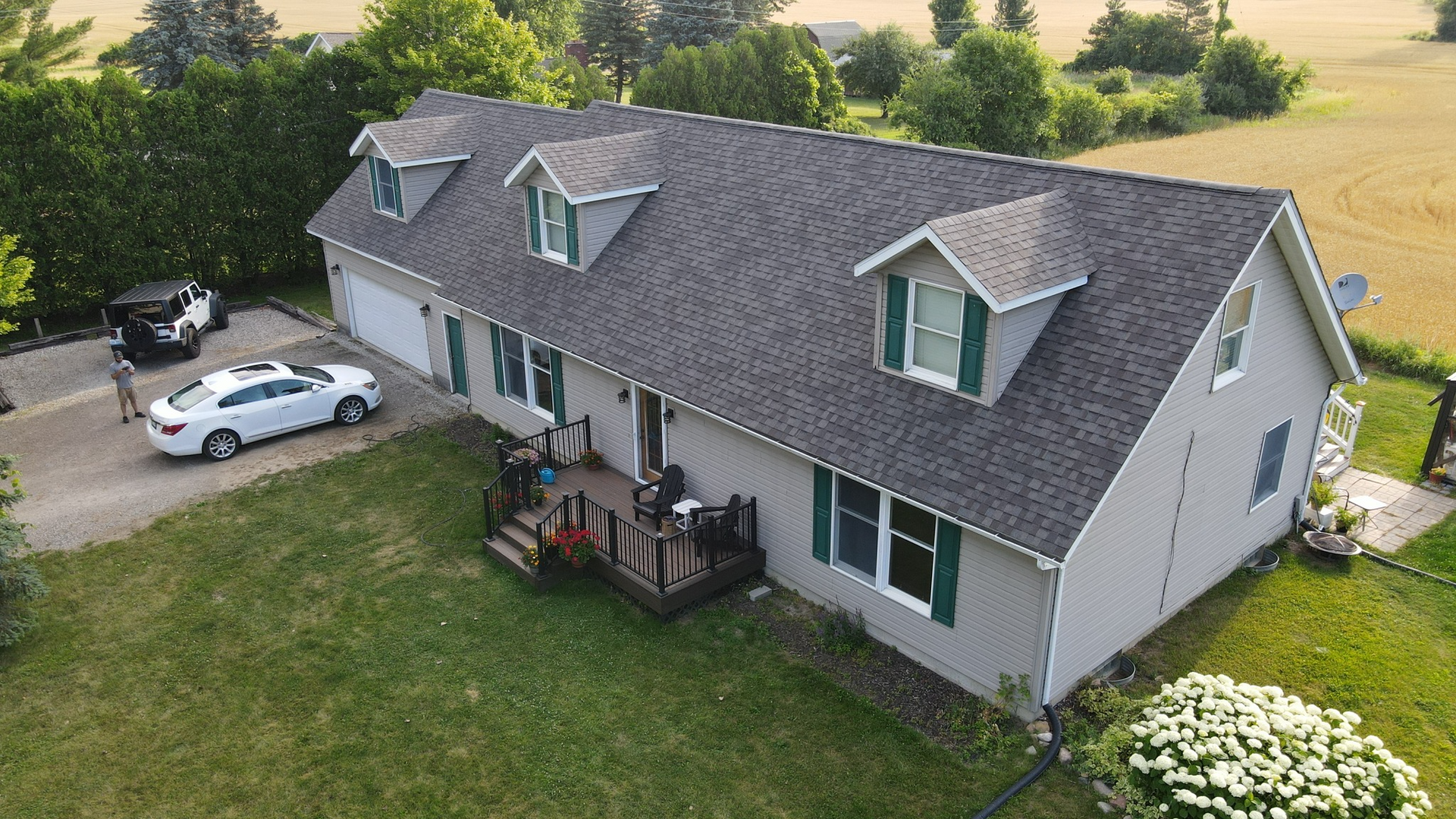 Gray house with three dormers, green shutters, and a dark deck; cars in the driveway and a field in the background.