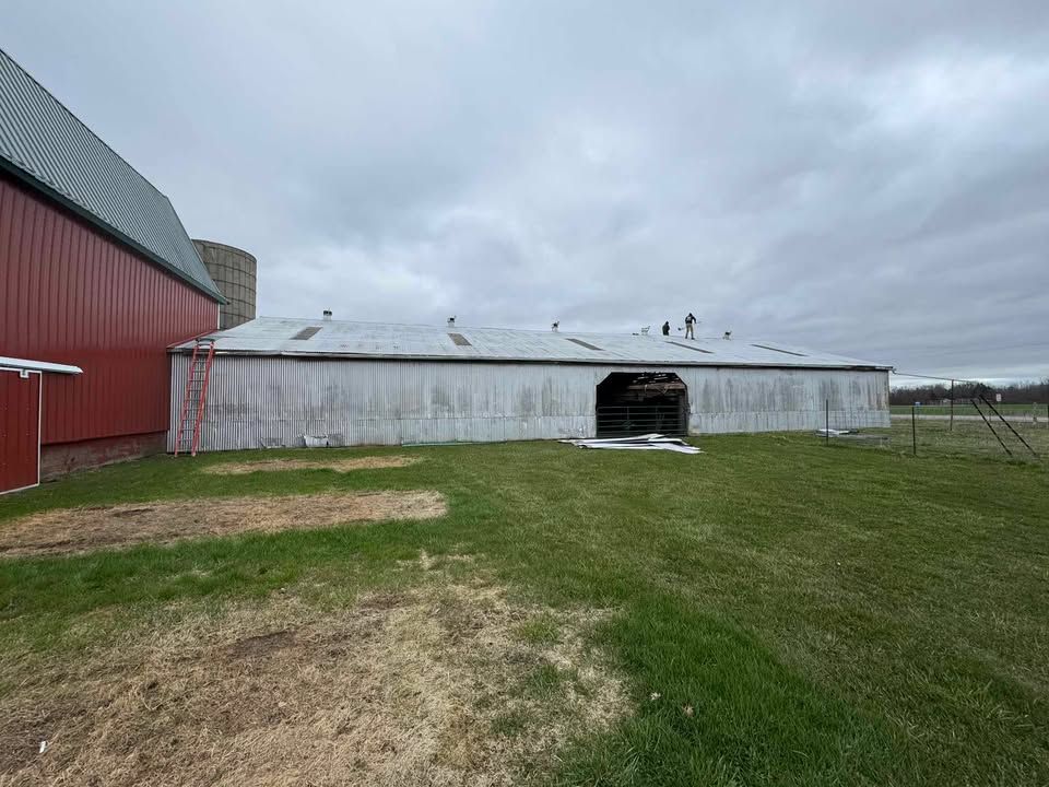 A long white barn with a partially collapsed roof and a red barn to the left, cloudy sky.