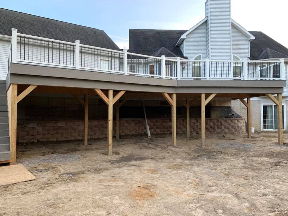 Elevated wooden deck attached to a light-colored house. Brown support beams and railing. Dirt ground.