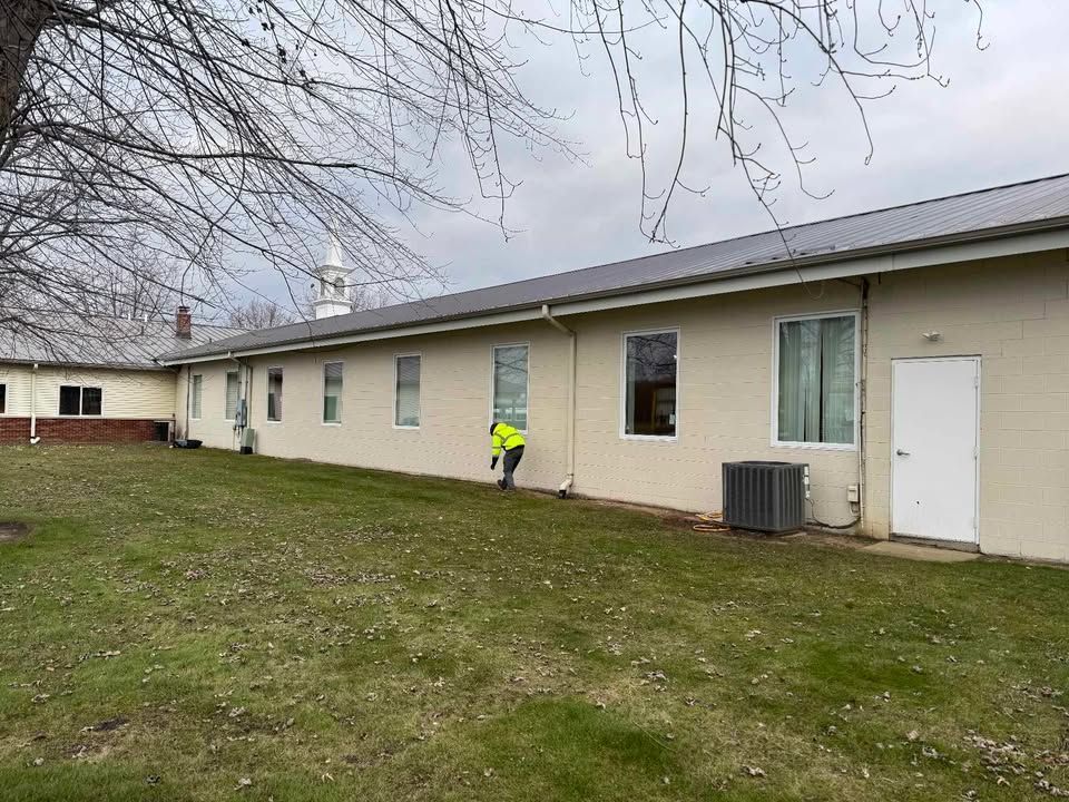 Person in safety vest sweeping beside a beige building with a metal roof. Overcast day.
