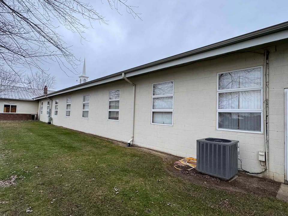 Exterior view of a beige building with multiple windows, a green lawn, and an AC unit. Cloudy sky.