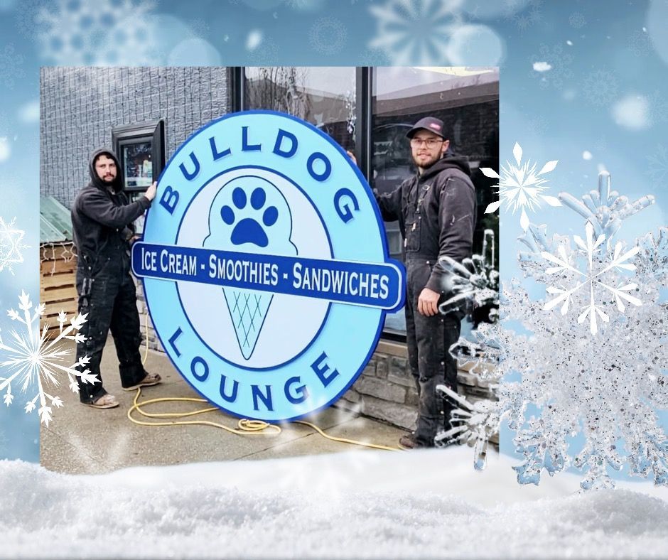 Two men installing a large, round Bulldog Lounge sign with ice cream cone, paw print, and blue text. Snowflakes surround the image.