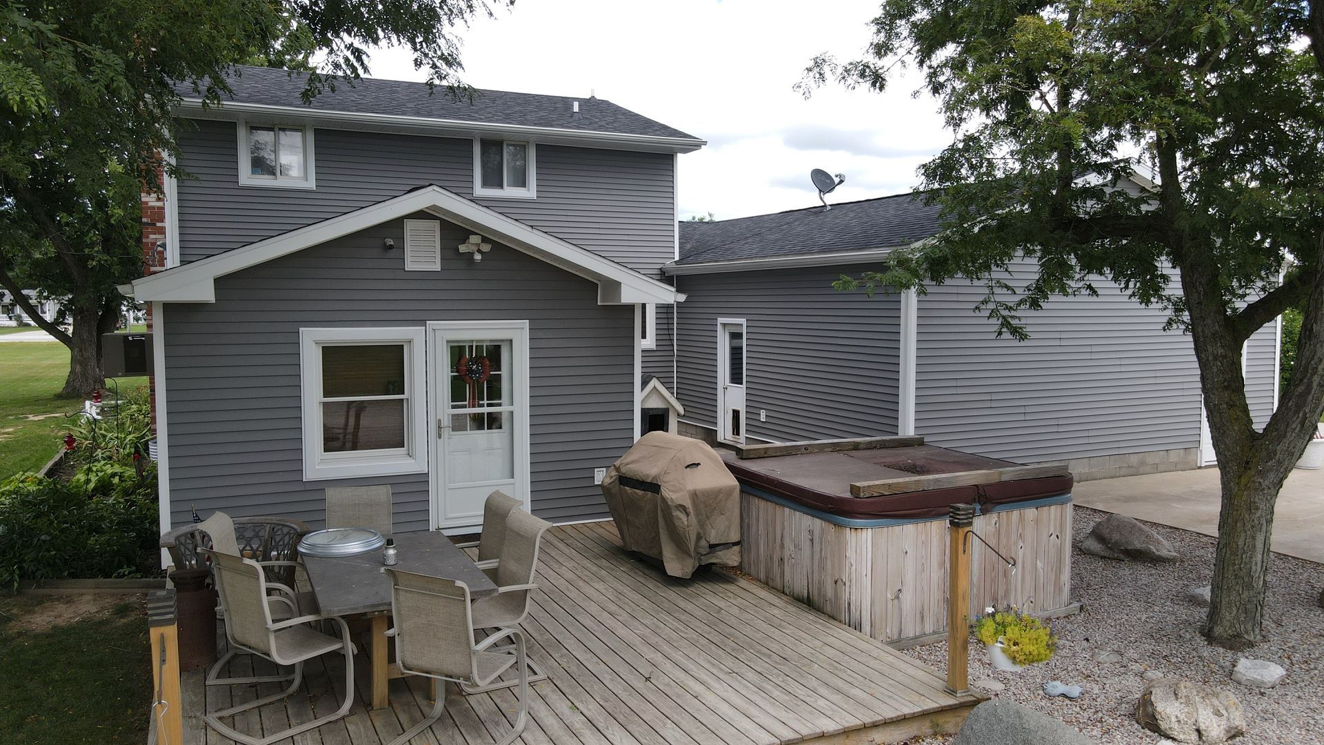Gray house with deck, table, chairs, grill, and hot tub. Tree on right.