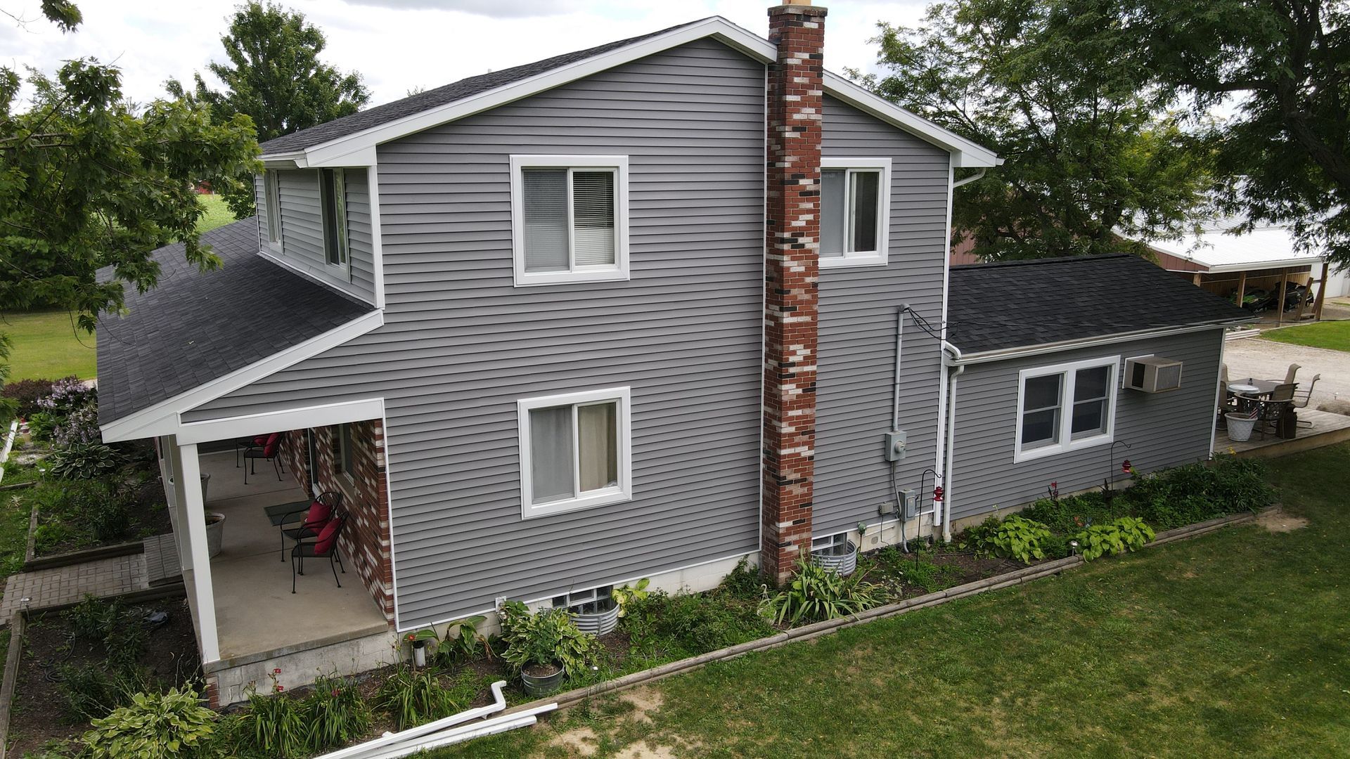 Gray-sided two-story house with a black roof, white-trimmed windows, and a brick chimney. Green lawn and trees surround the home.