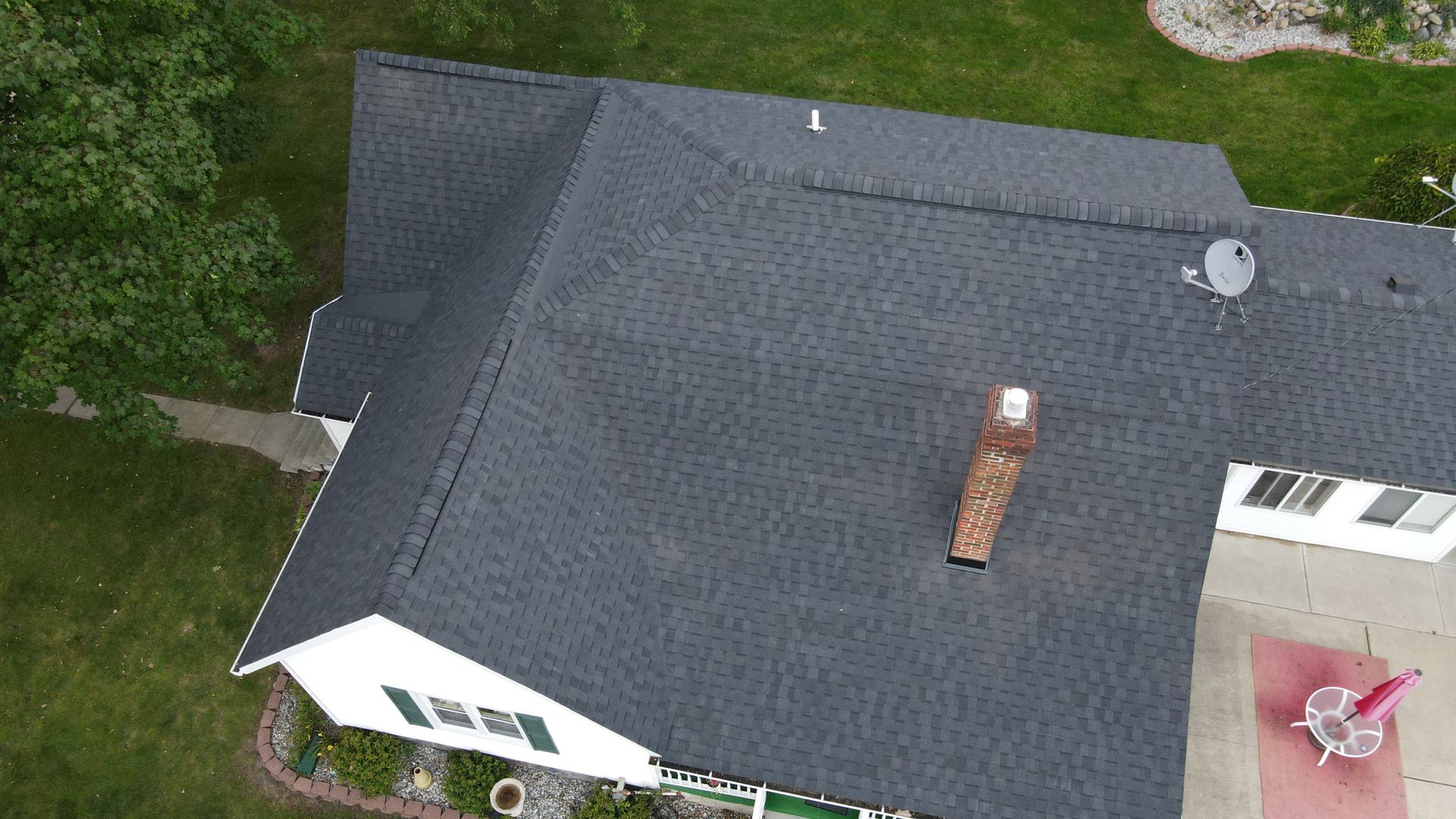 Overhead view of a dark gray shingled roof with a chimney, a satellite dish, and a white house with a patio.