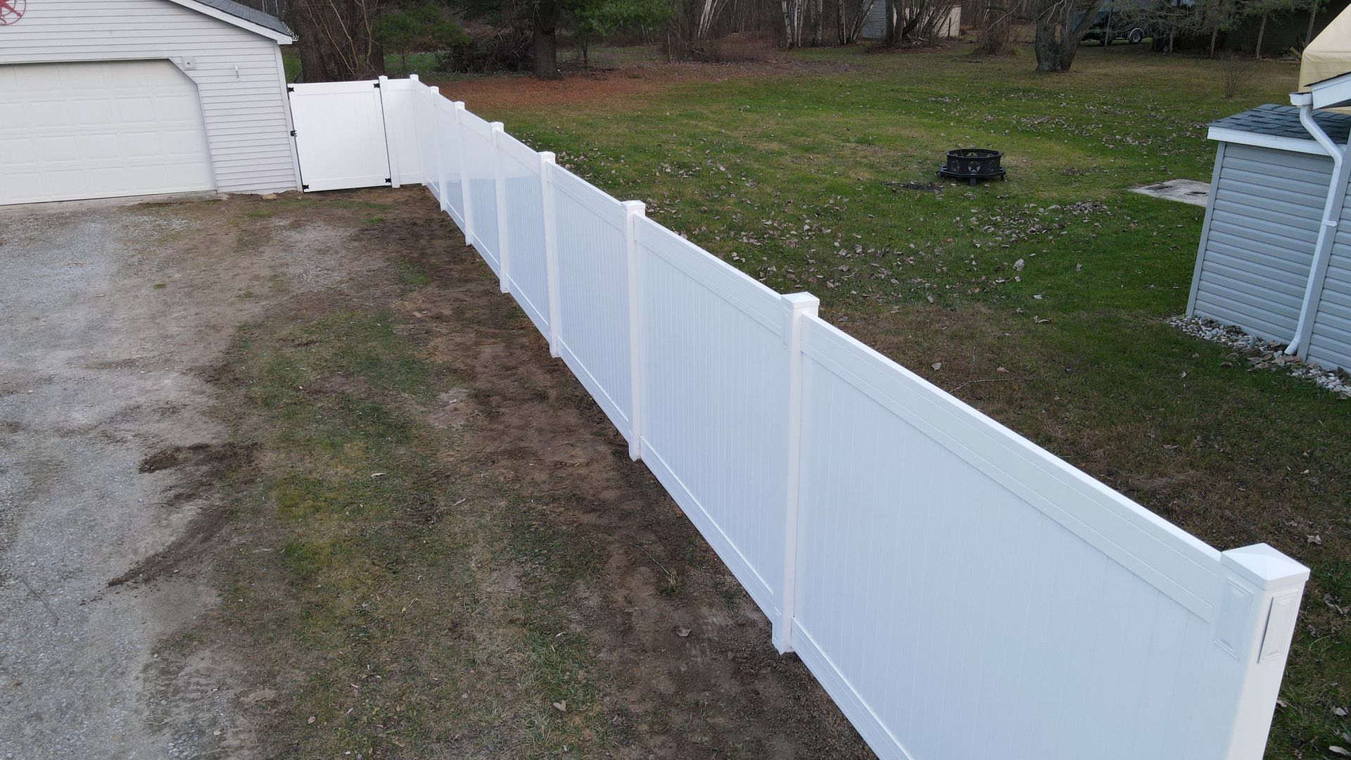 White vinyl fence bordering a gravel driveway and grassy yard, with a shed and garage visible.