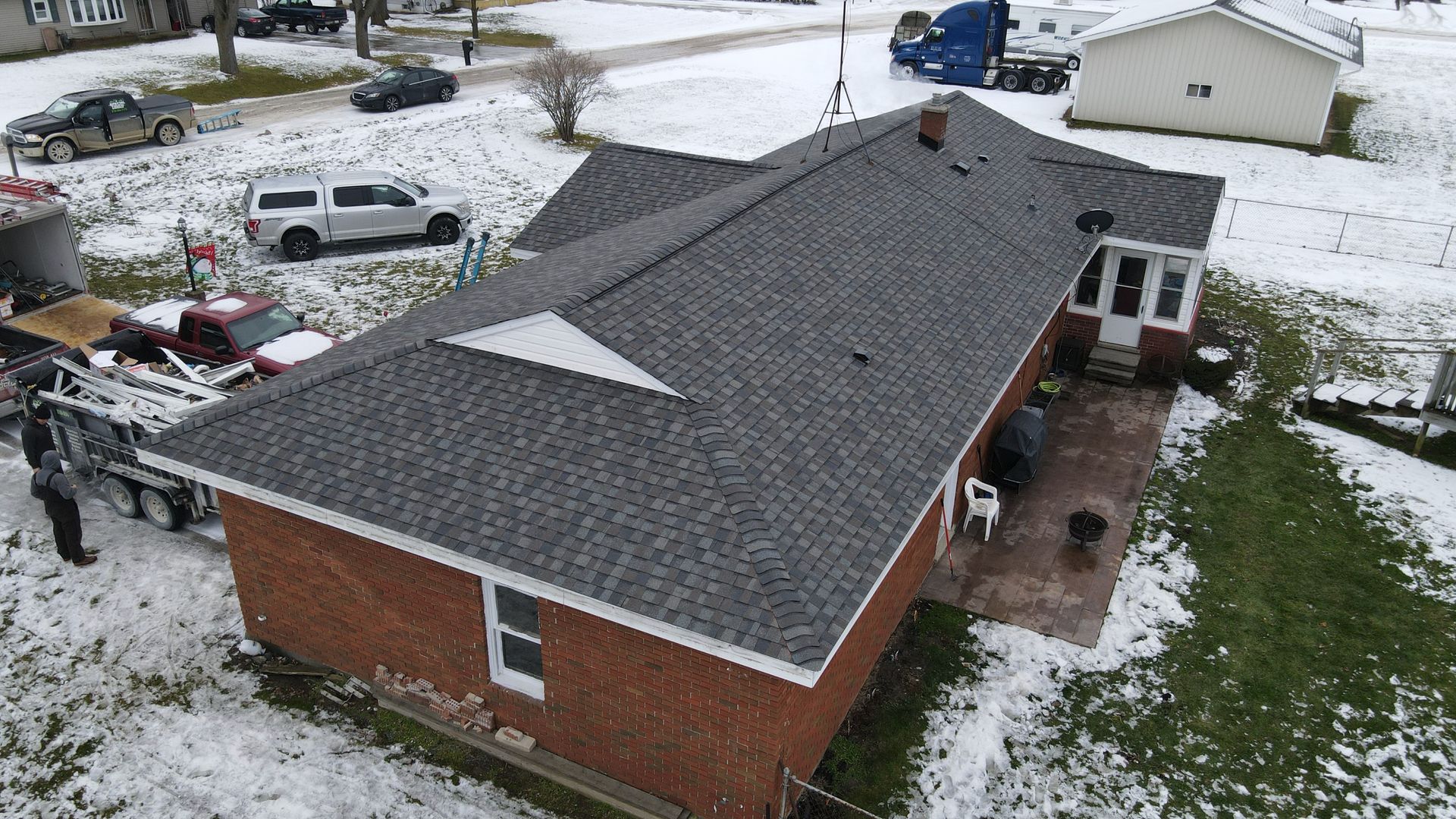 Aerial view of a brick house with a newly installed gray shingle roof, surrounded by snow.