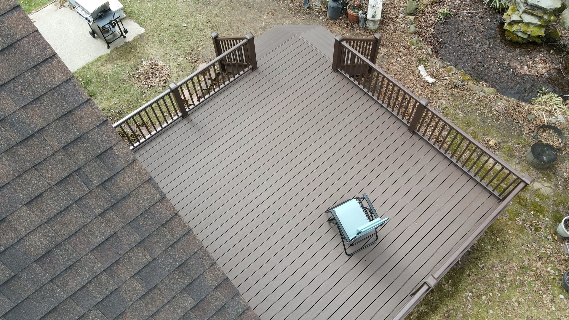 Overhead view of a brown wooden deck with a chair, next to a brown roof, and surrounded by grass.