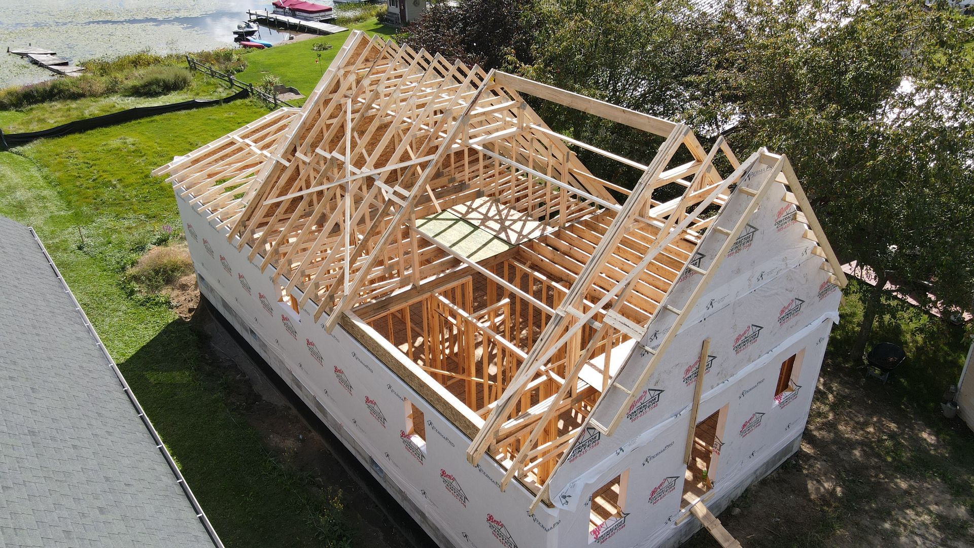 Wood-framed house under construction; exposed rafters, sheathing; setting outdoors, near water.