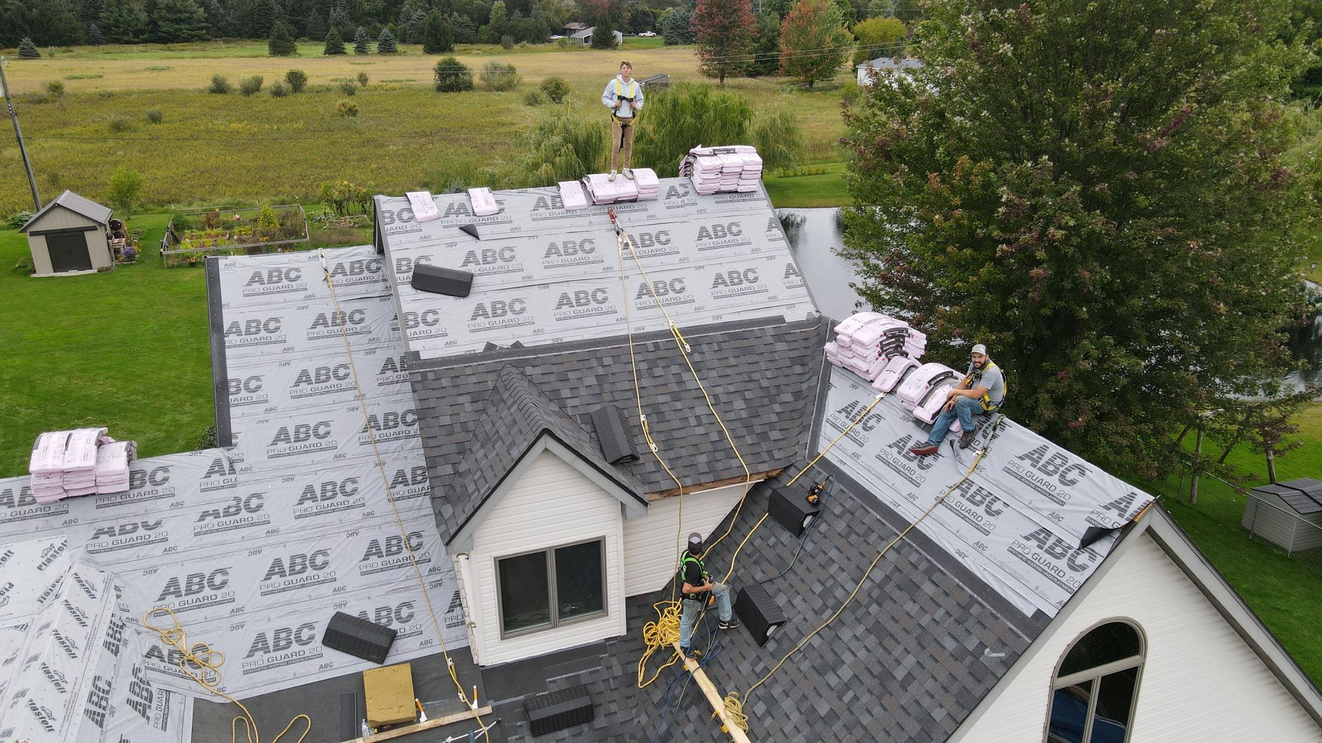 Roofers installing shingles on a house with a green lawn and trees in the background.