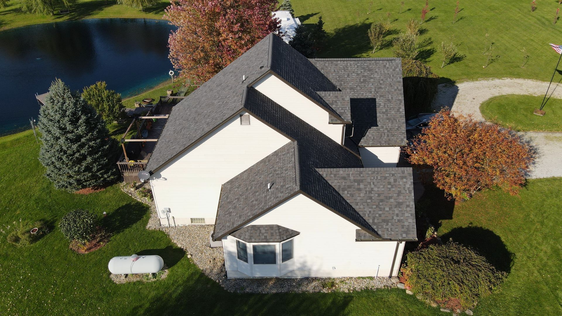 Aerial view of a white house with a dark gray roof, surrounded by green grass, a pond, and trees.