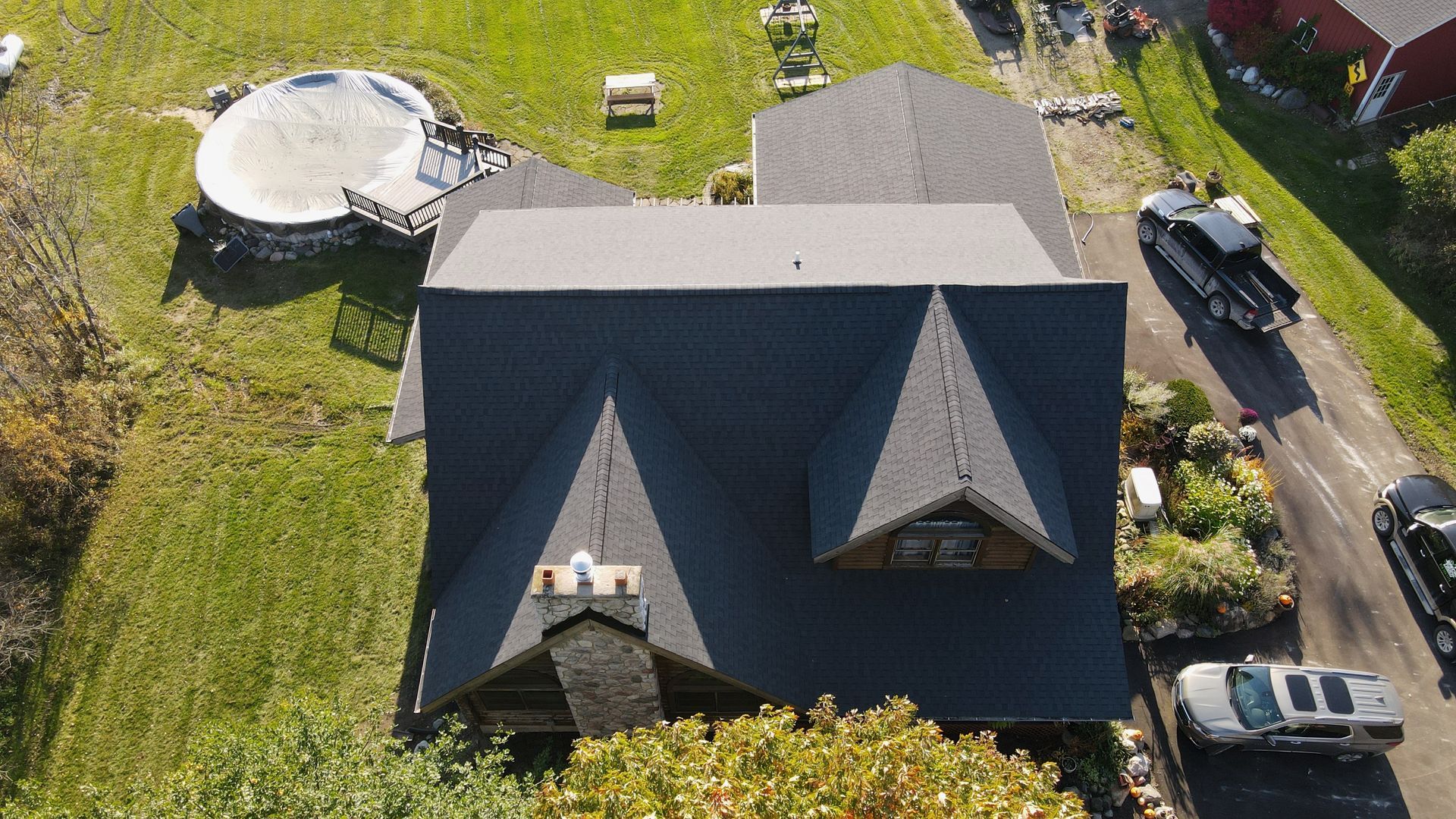 Overhead view of a house with a dark gray roof, chimney, and vehicles in a driveway. A round patio is in the distance.
