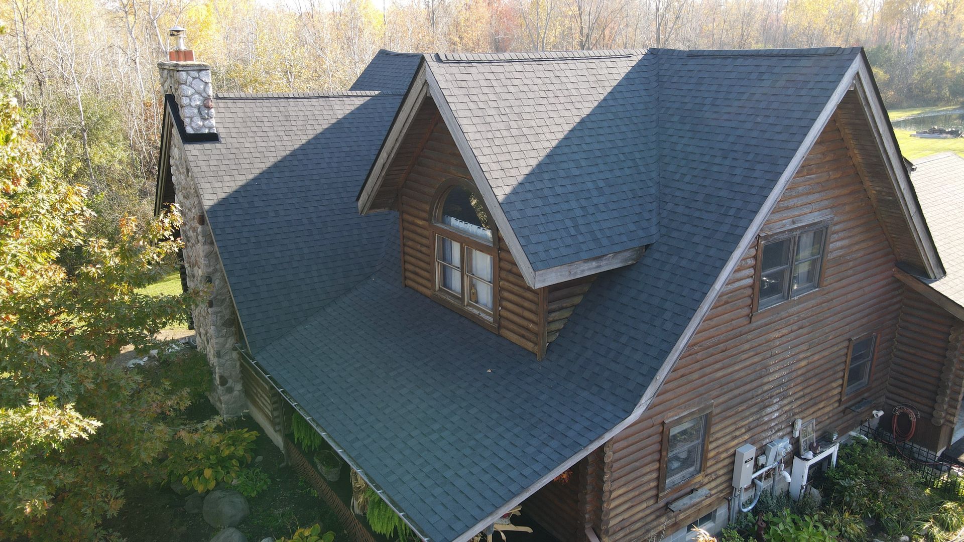 Log cabin with dark gray roof, two dormers, and chimney surrounded by trees.