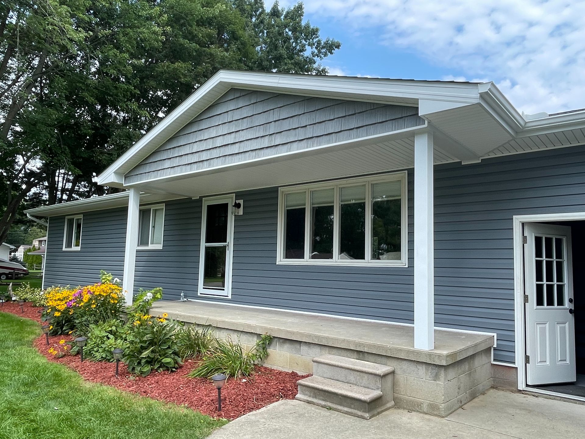 Blue-sided house with a porch and white trim.  Landscaping includes red mulch and yellow flowers.