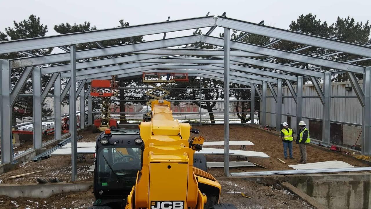 Construction site of a metal building frame. Yellow JCB telehandler in foreground. Two workers stand inside.