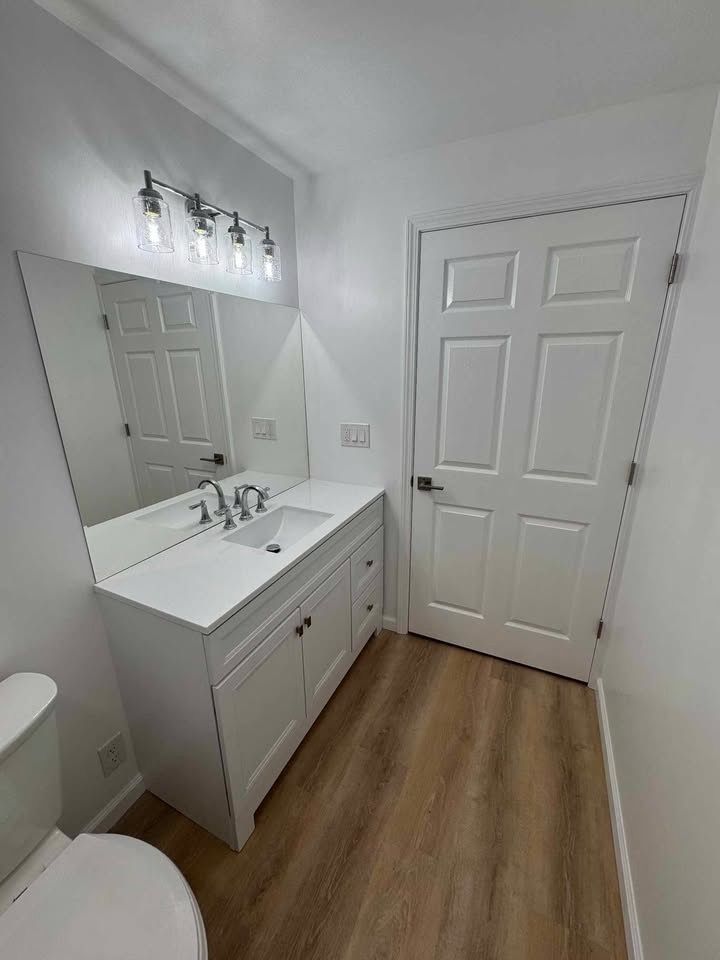 White bathroom with vanity, sink, mirror, door, and wood-look flooring.