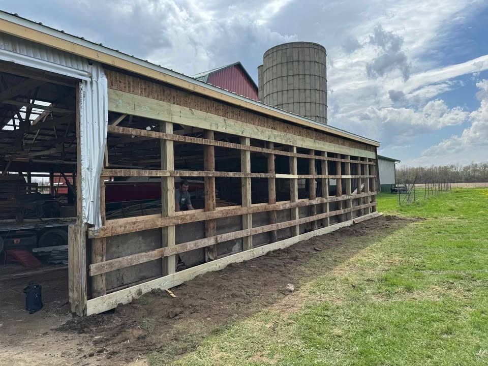 Barn with open wall framing, construction in progress, silo in background, cloudy sky.