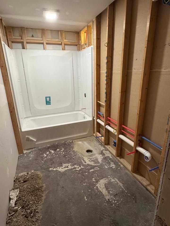 Bathroom renovation: framed walls, white tub/shower, exposed plumbing, concrete floor with debris.