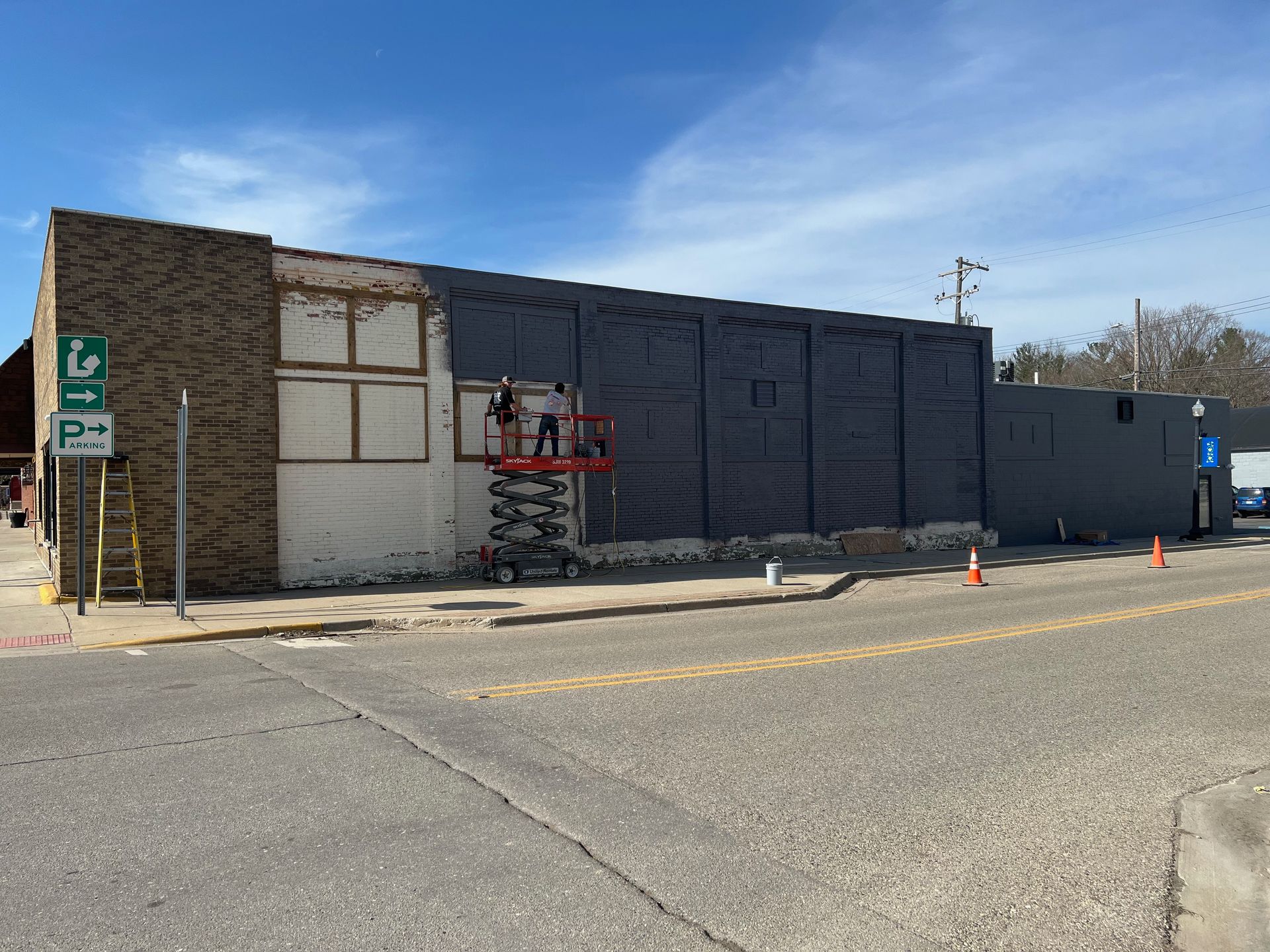Building exterior under renovation. Workers on lift applying dark siding. Street and blue sky in background.