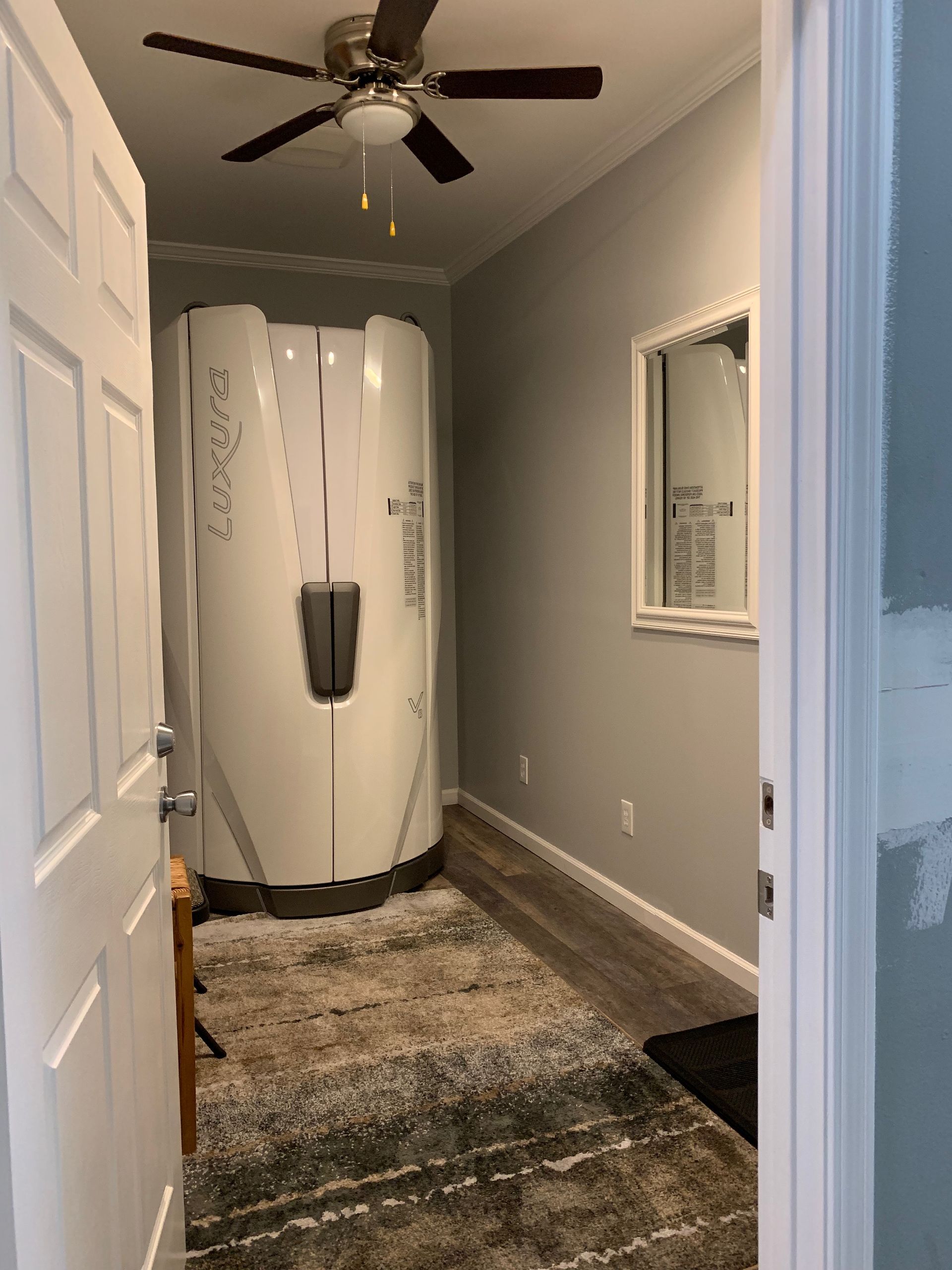 Room with tanning bed, mirror, and rug. Doorway to the left and ceiling fan overhead. Gray walls.