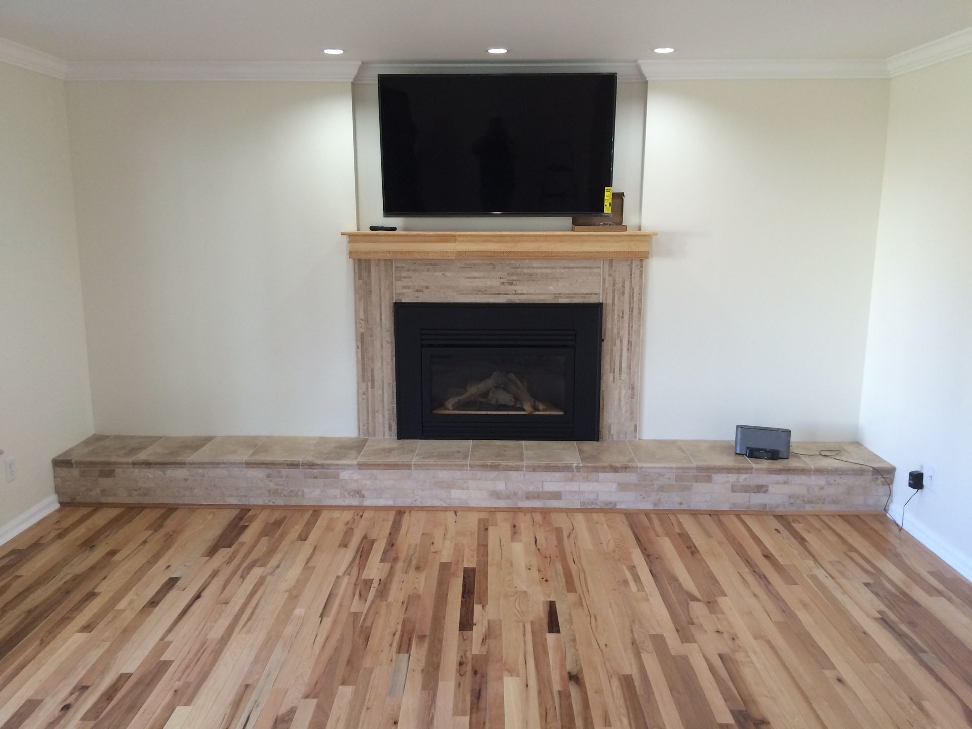 Fireplace with mounted TV, light-colored stone and wood trim. Light wood flooring. Recessed ceiling lights.
