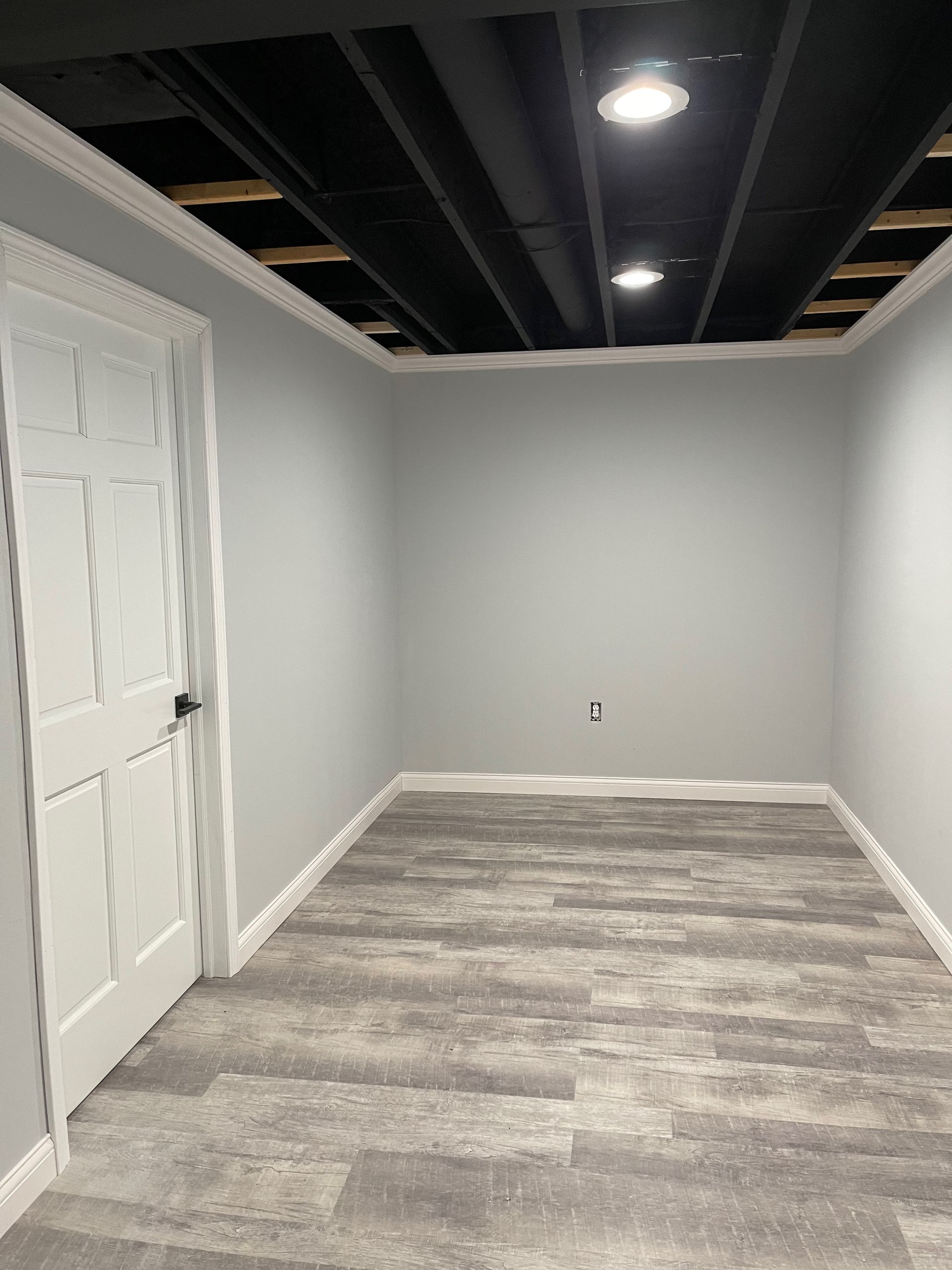 Empty room with gray walls, wood-look flooring, white trim, door, and a partially open black ceiling with recessed lights.