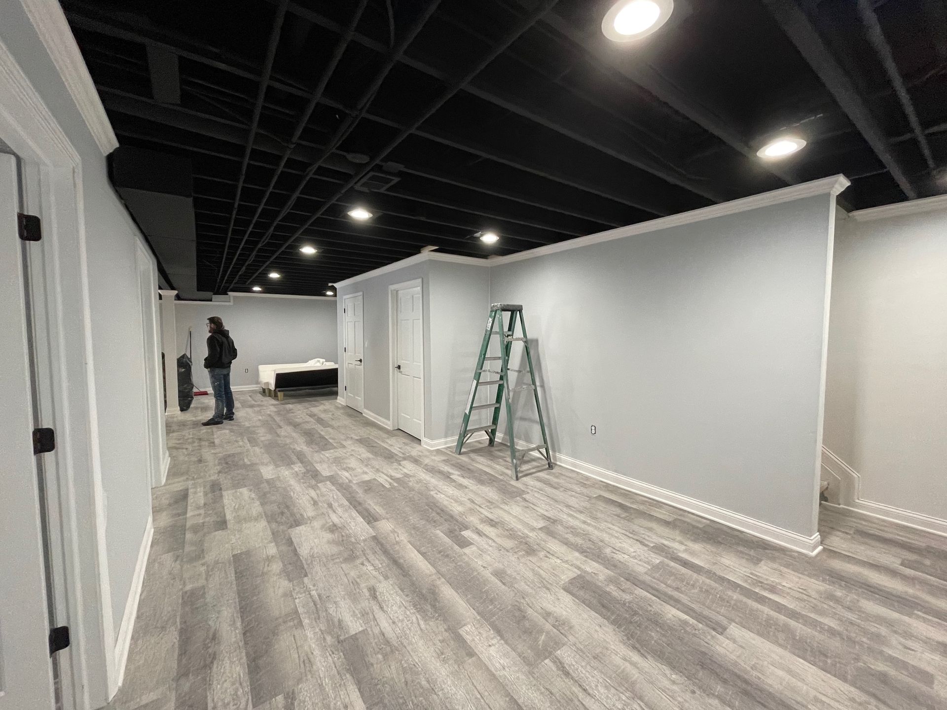 Basement renovation with gray floors, black ceiling, white trim, and a person standing near a wall.