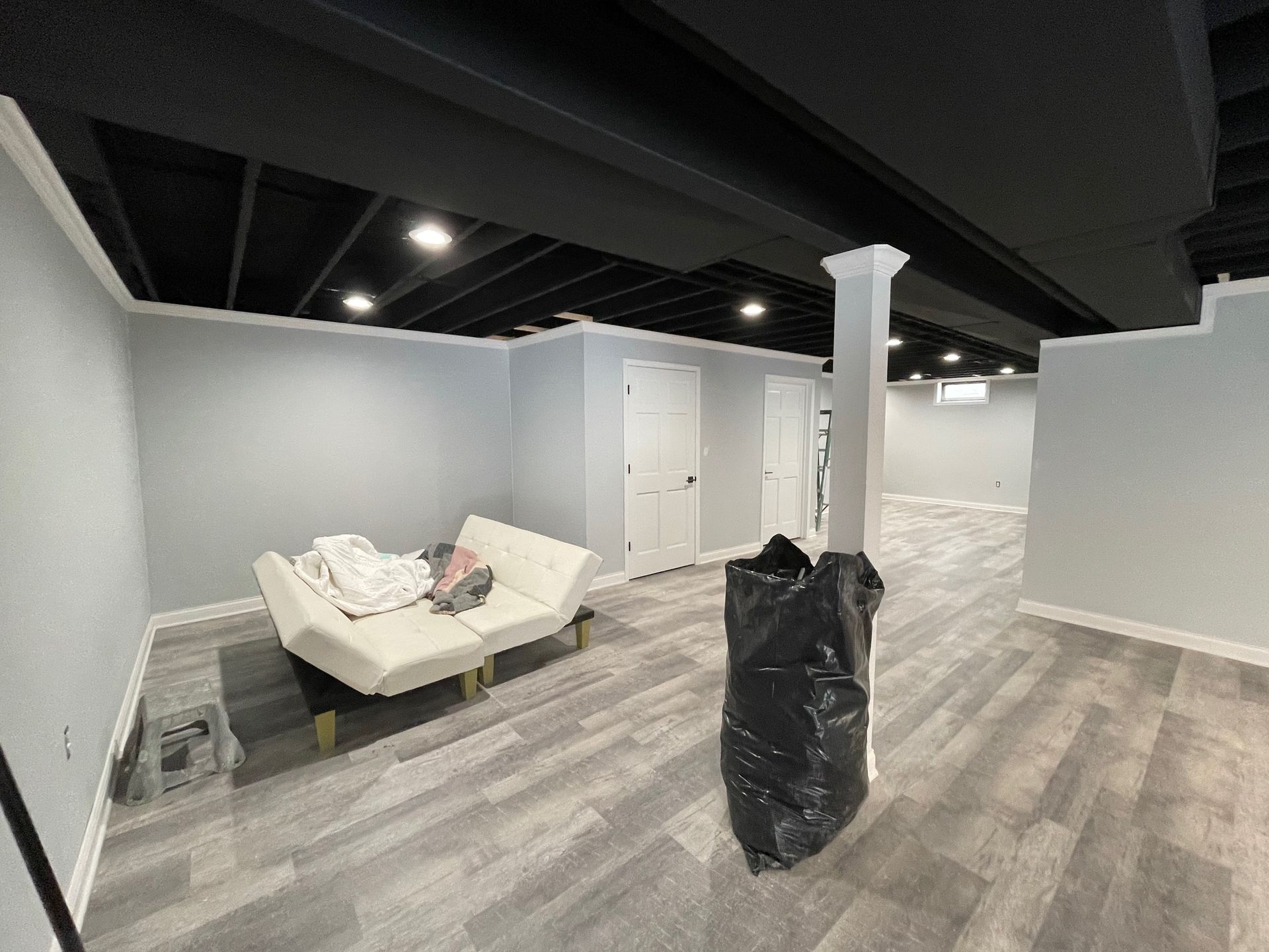 Finished basement with light gray walls, black ceiling, white trim, and gray flooring. A white couch, doors, and a support column are present.