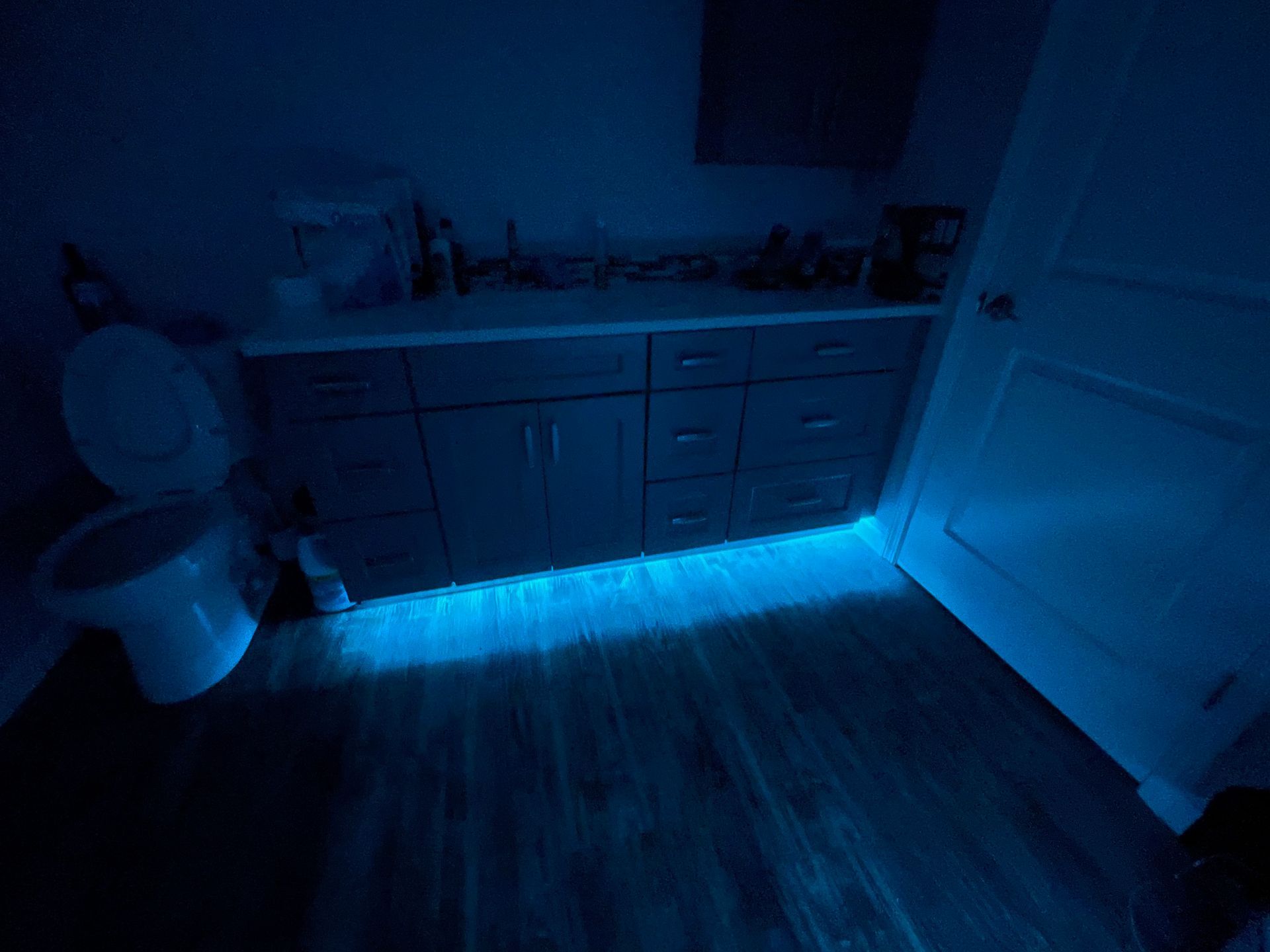 Blue LED strip lights illuminate a bathroom floor. A toilet, vanity, and open door are visible.