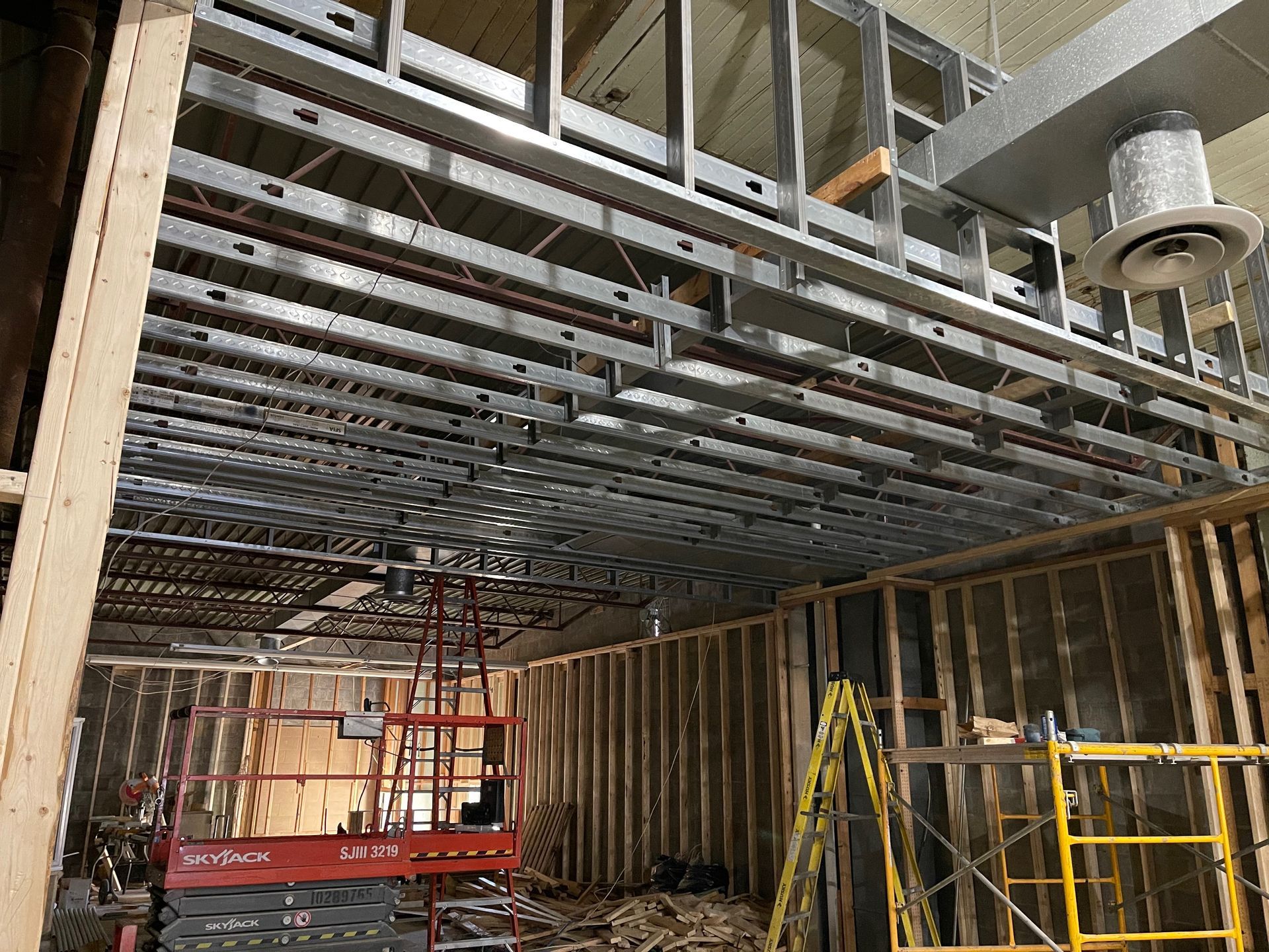 Construction site interior. Metal ceiling framing installed, wooden walls being built, lift and scaffolding present.