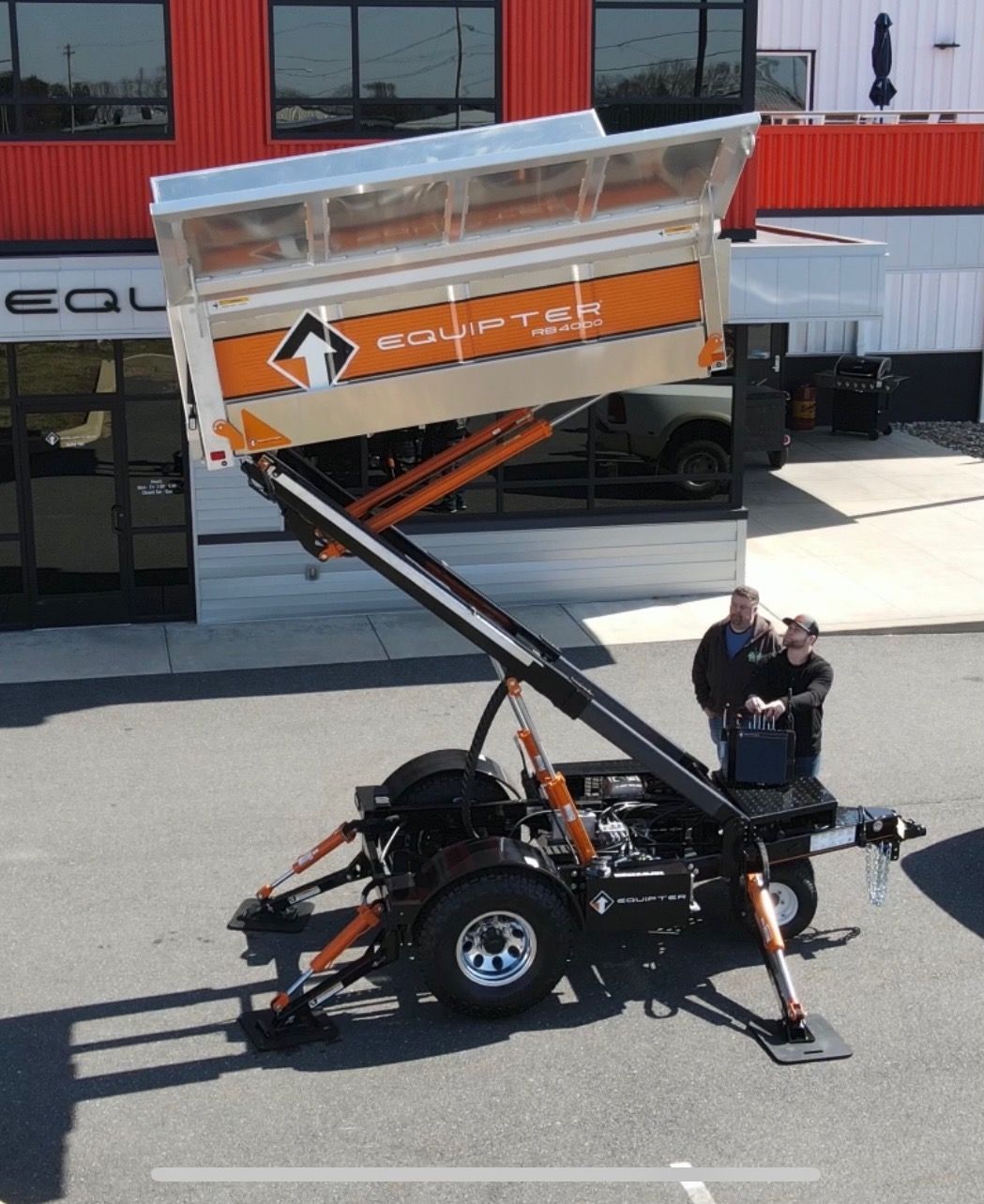 A raised orange and silver dumpster on a trailer. Two men observe. Red building in background.