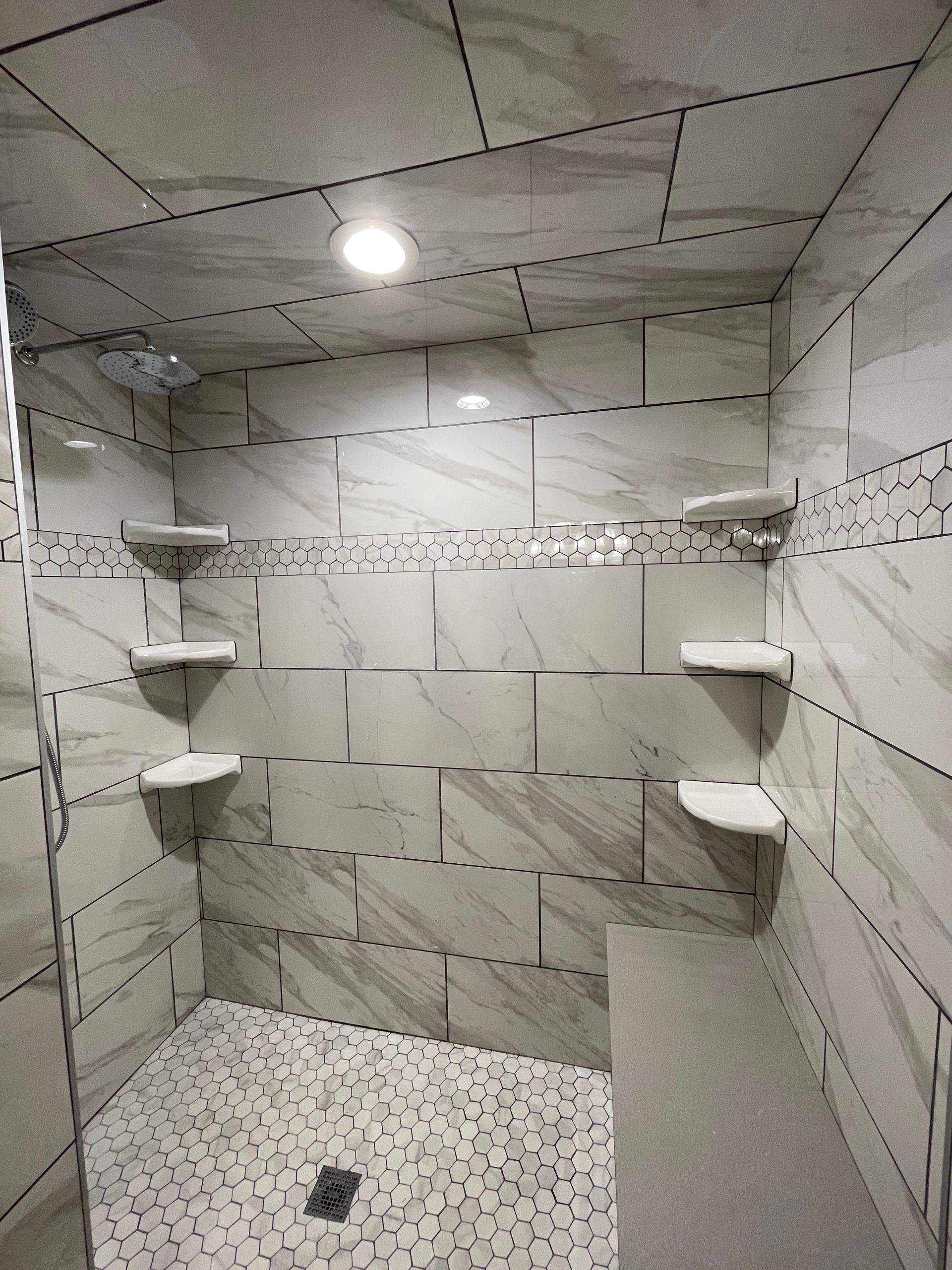 A tiled shower with built-in shelves, pebble floor, and a bench. White and gray tones, overhead light.