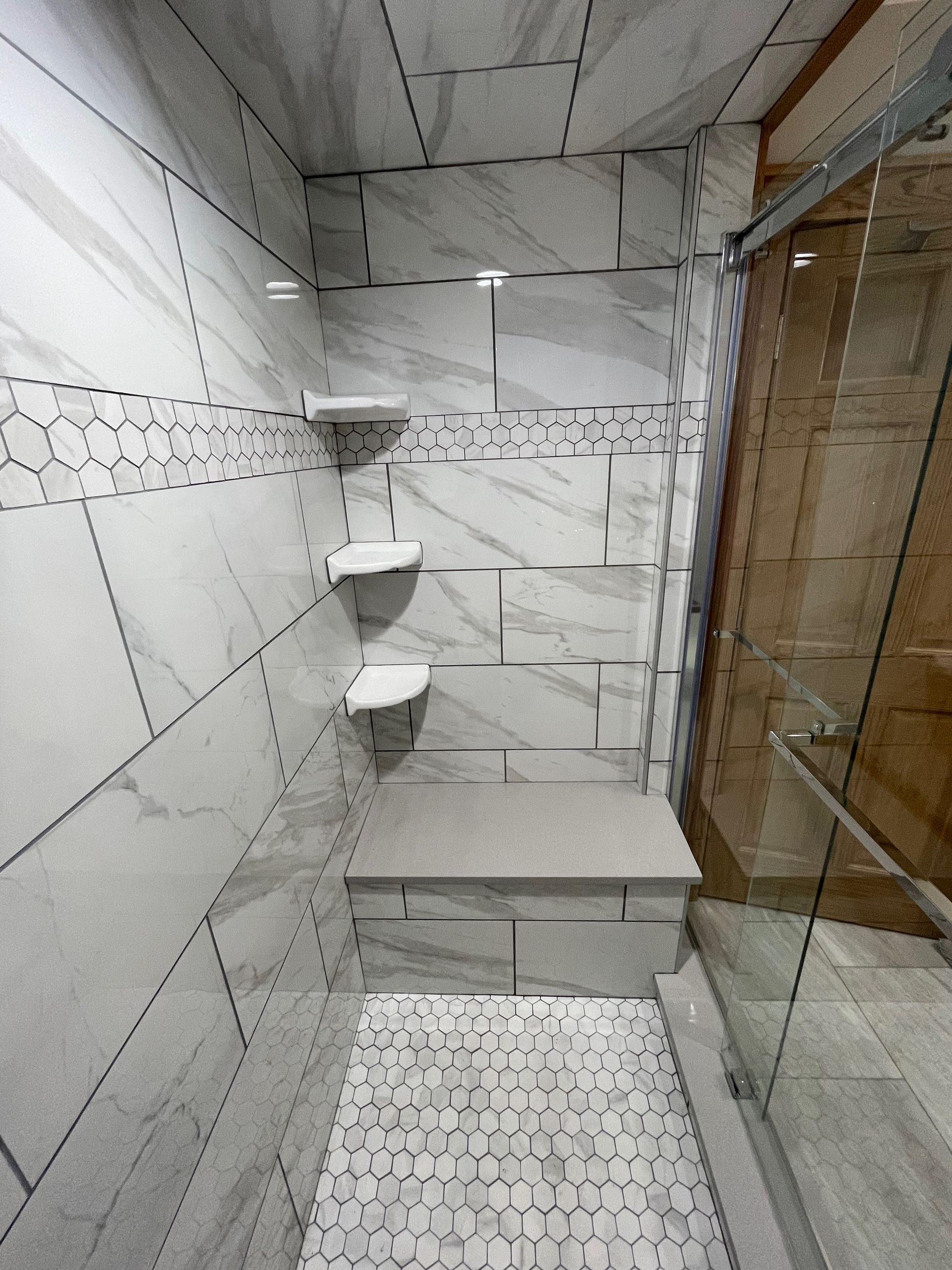Tiled shower with built-in shelves and bench. Glass door, white and gray tiles.