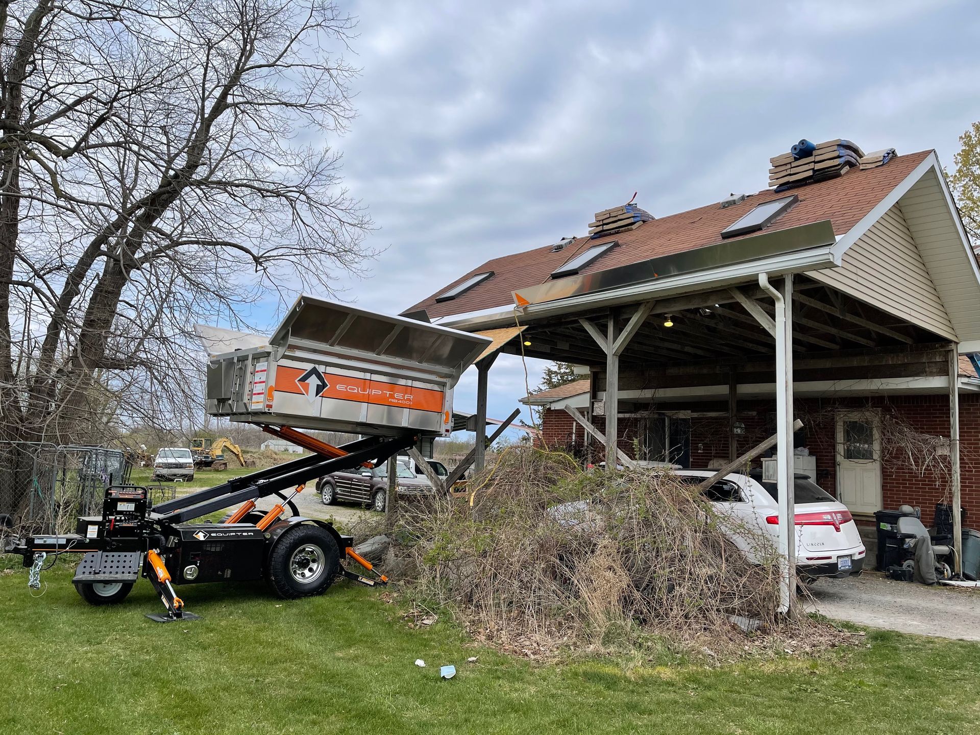 Roofing debris removal. Orange/black lift truck beside a house with roof replacement in progress, on a cloudy day.